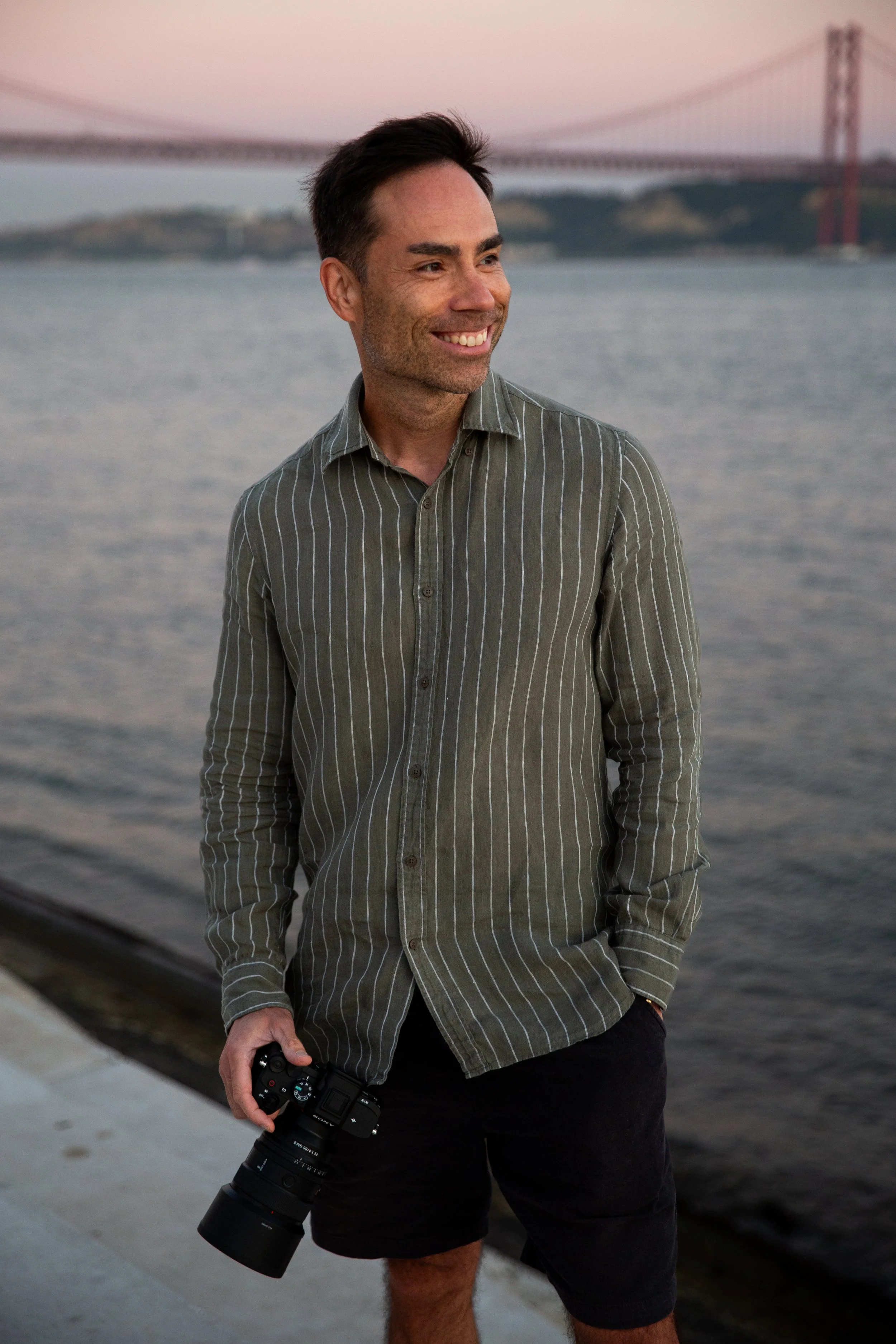 Portrait of a photographer holding his camera, smiling by the Tagus River, with the 25 de Abril bridge in the background in Lisbon.