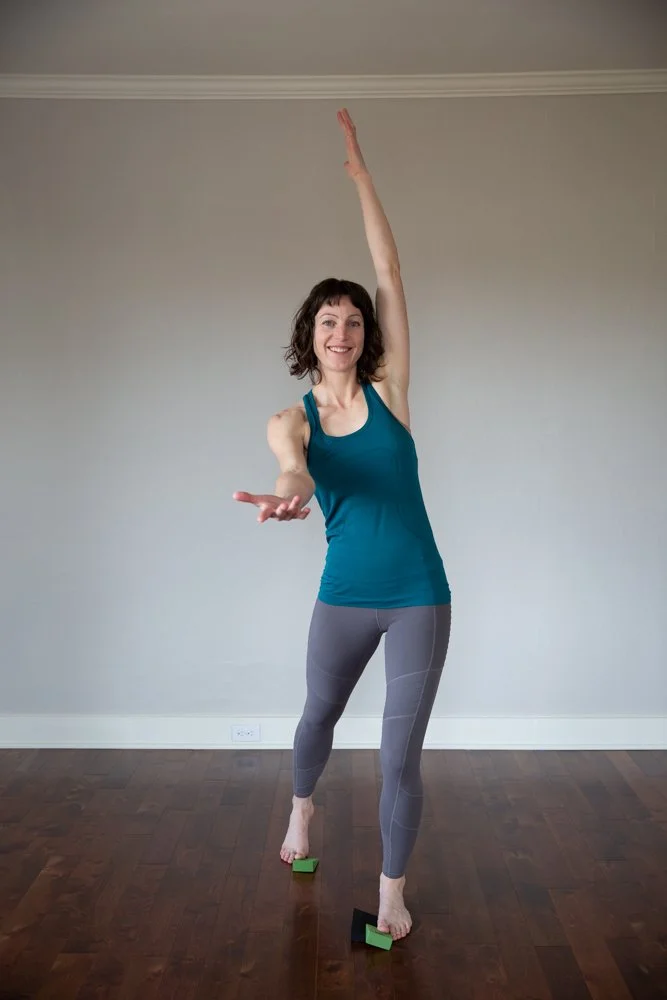 A woman in workout clothes balancing on two blocks with one arm raised high and the other extended in front, smiling, in a room with hardwood floor, during a personal branding photoshoot in Lisbon.