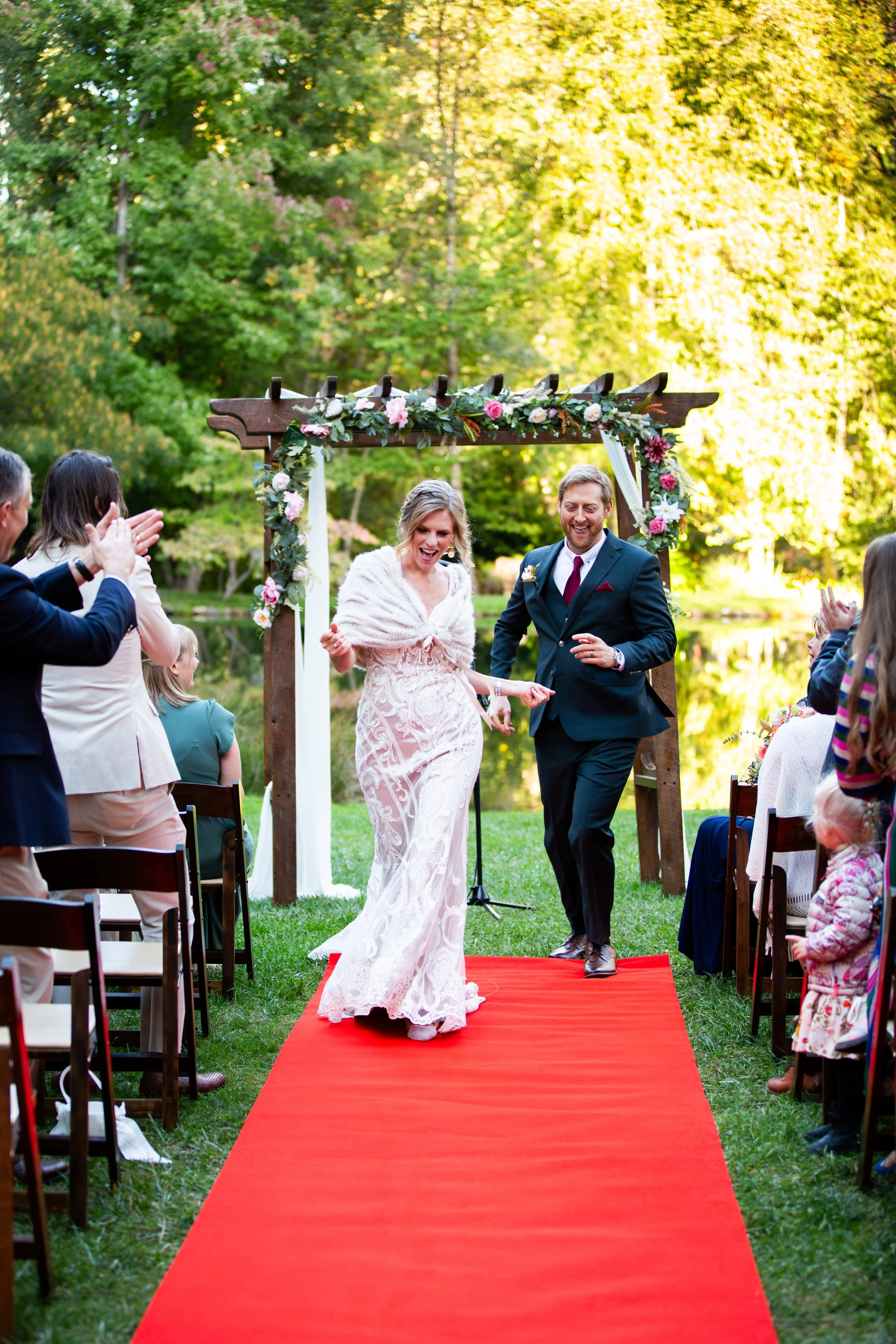 A bride and groom walking down a red carpet aisle under a wooden arch decorated with flowers at an outdoor wedding ceremony surrounded by guests enjoying the event, with lush green trees in the background in Nevada.