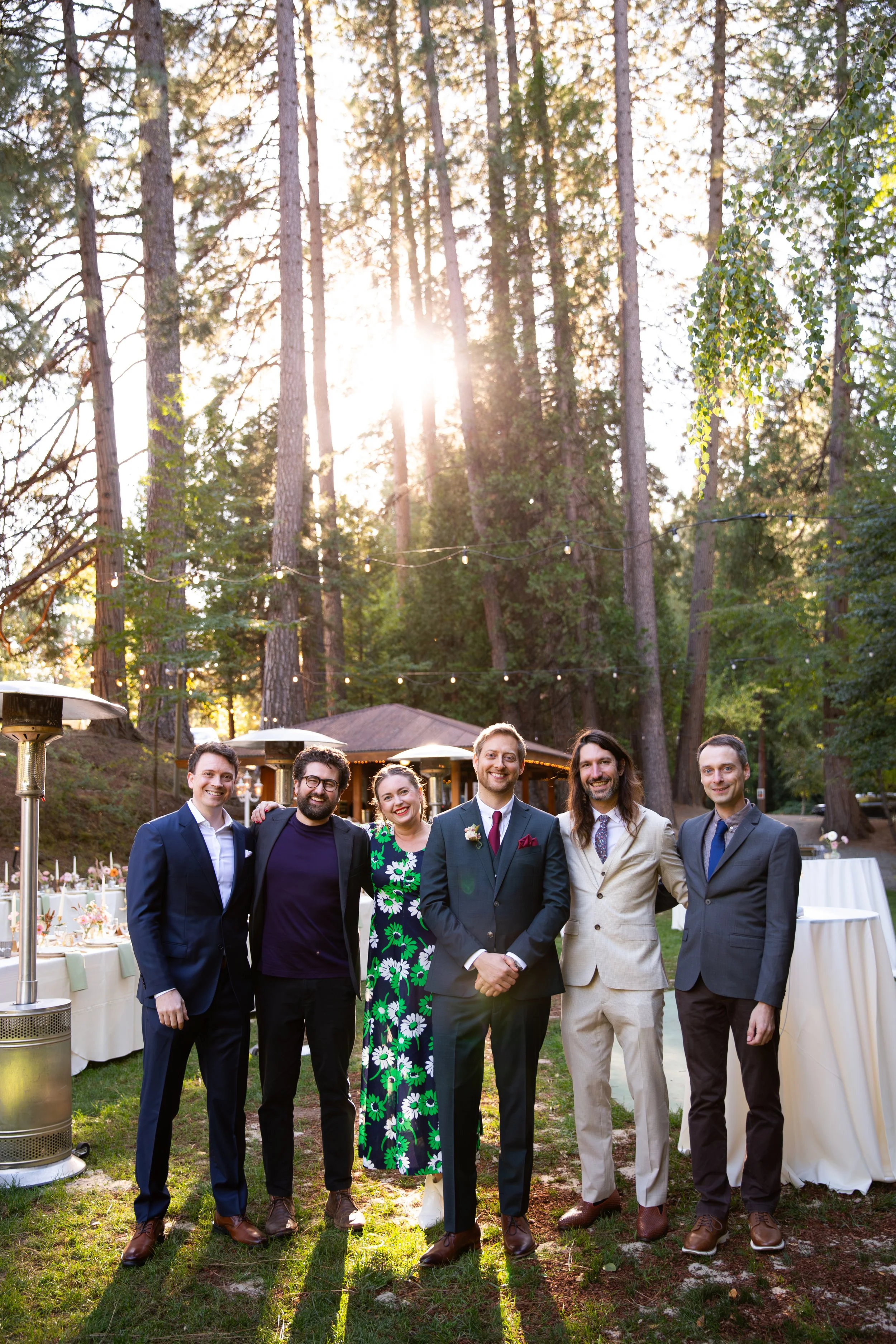 Group of six groomsmen and a groom dressed in formal attire standing outdoors at a wedding reception during sunset, with trees and string lights in the background in Nevada.