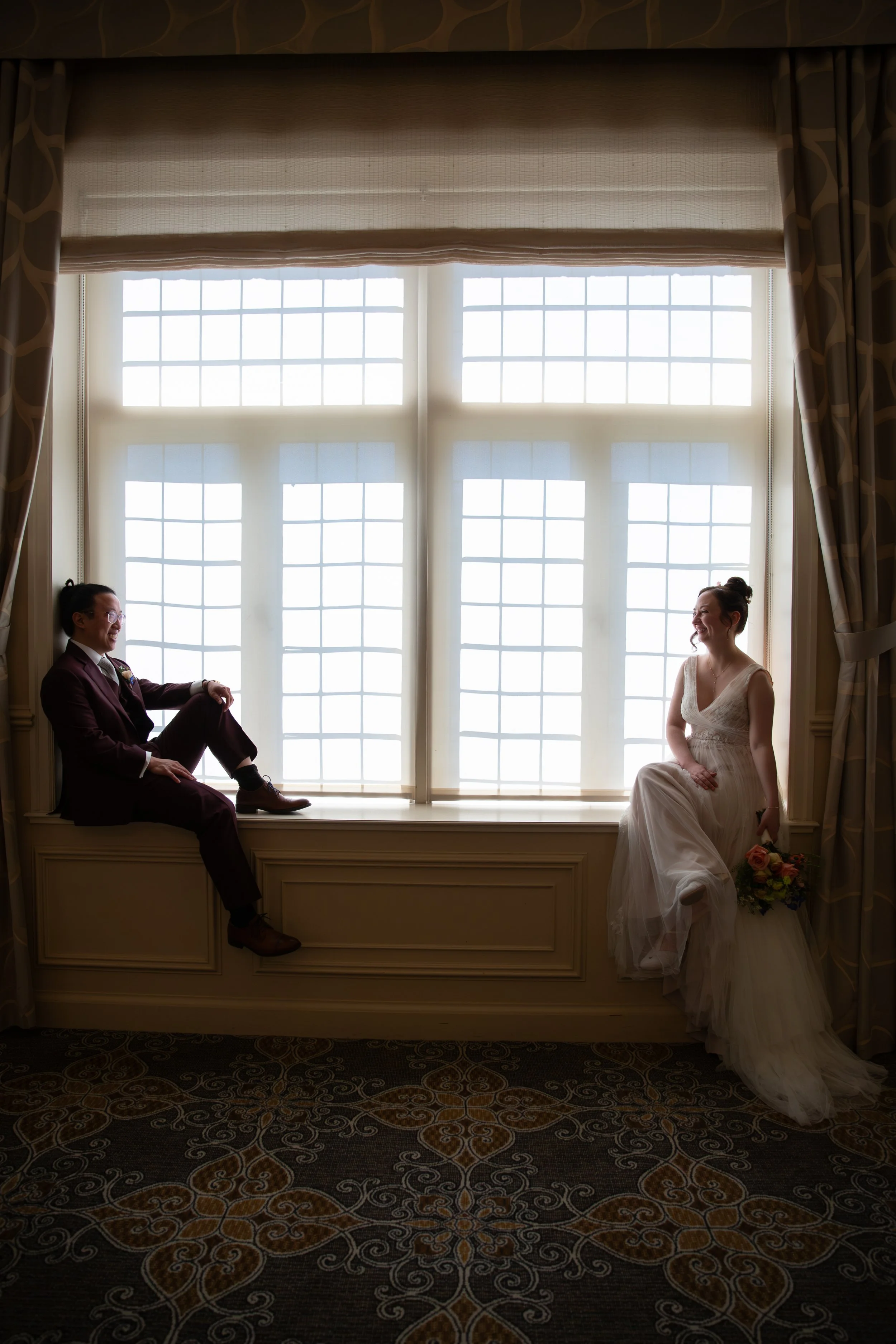 A bride in a wedding dress and a groom in a suit sitting and smiling inside a room with large window panes behind them in Lisbon.