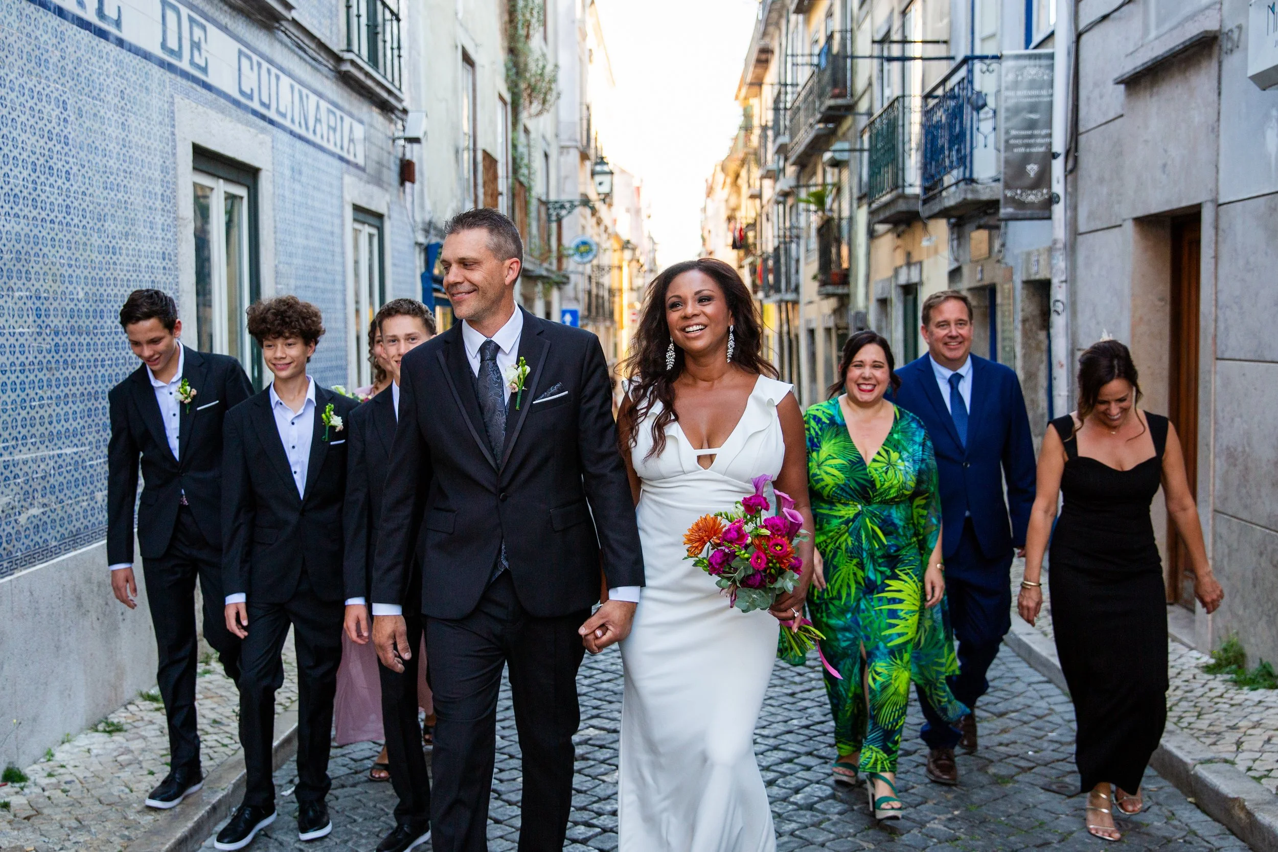 Wedding party walking on a small street in Lisbon while the bride and groom hold hands smiling in the front of the group. 