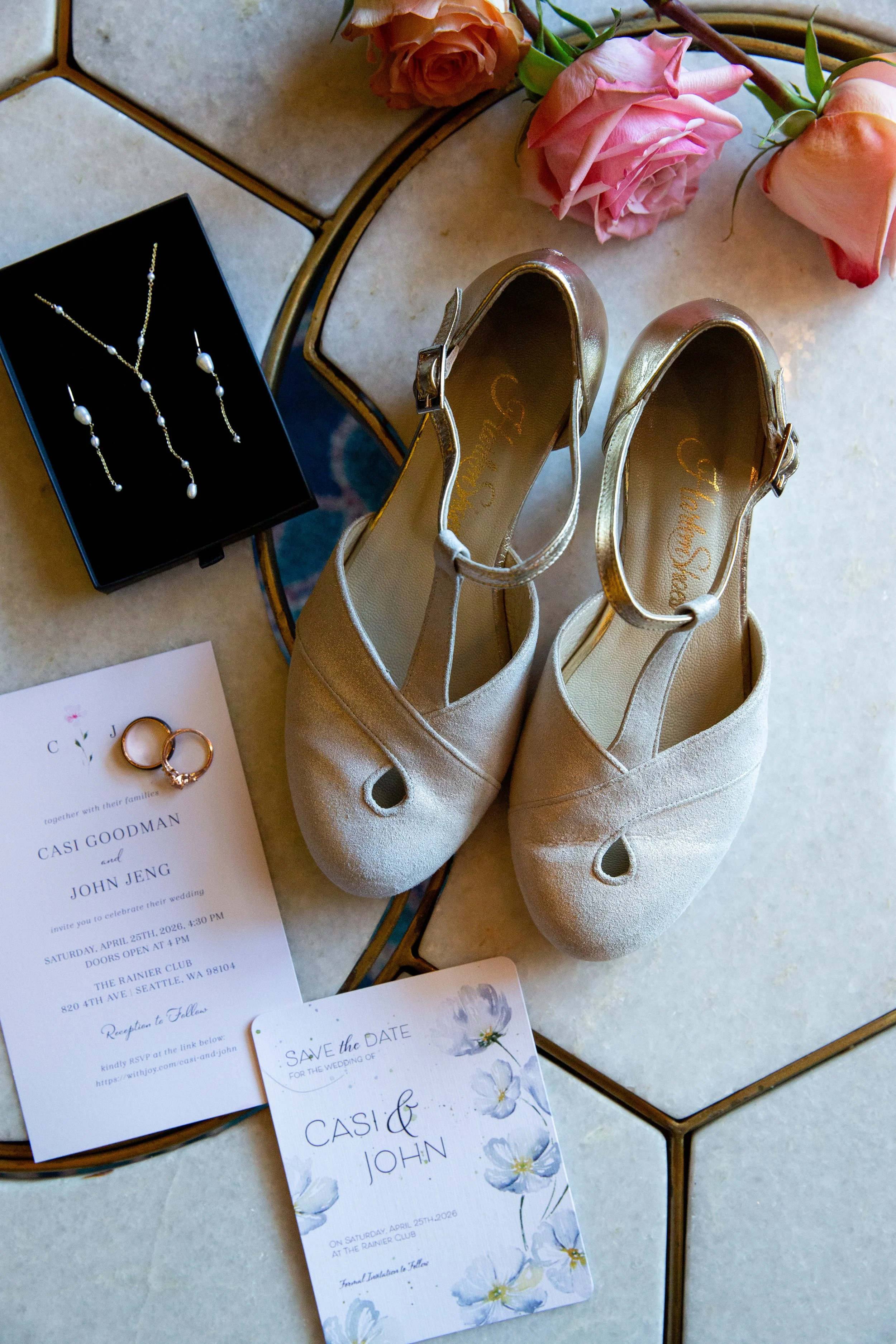 A pair of beige high-heeled shoes, a wedding invitation, a 'Save the Date' card, a jewelry box with pearl earrings and a necklace, a gold wedding band, and a few pink and peach roses on a decorative surface for a wedding detail photo in Lisbon.
