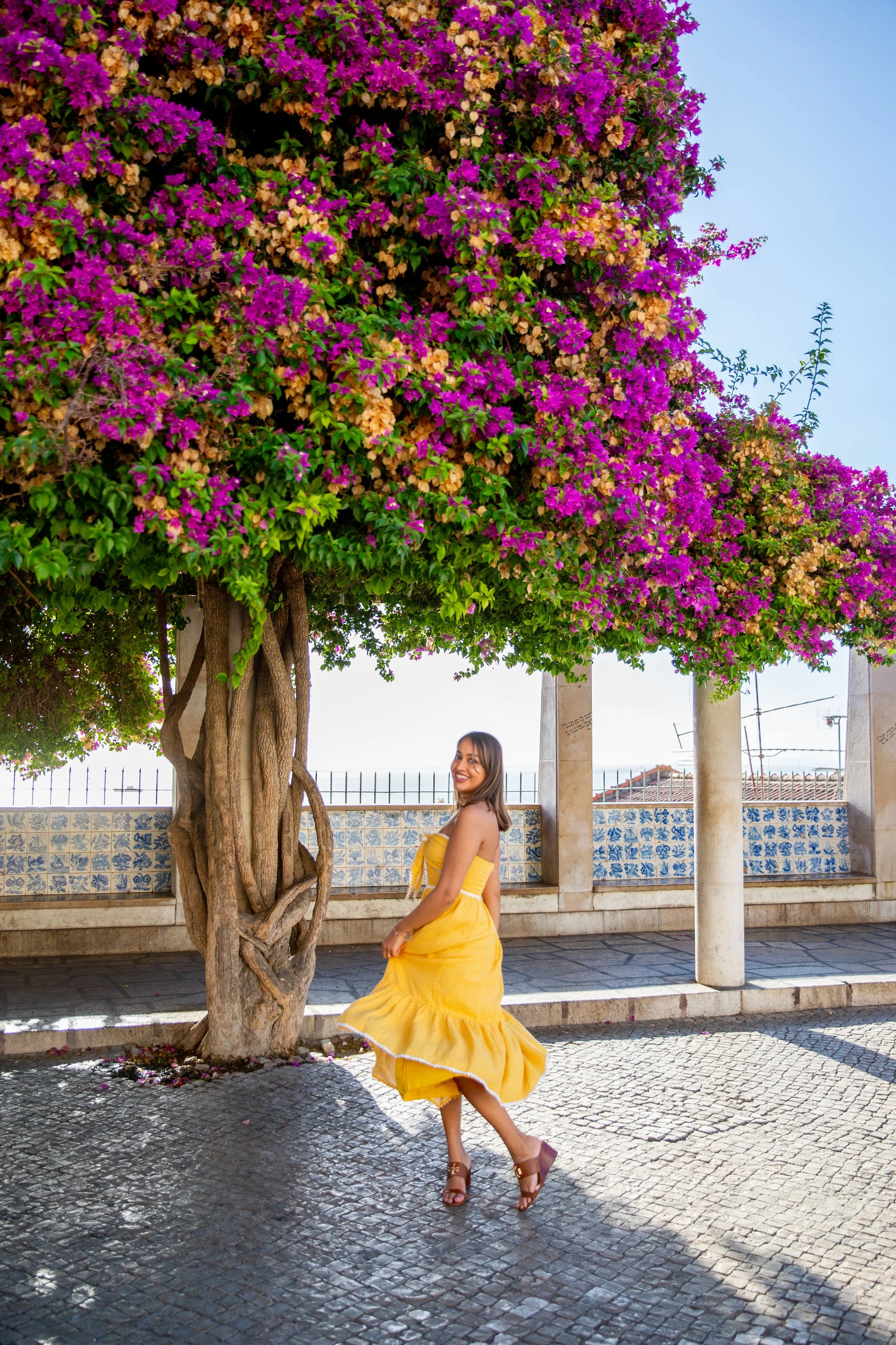 A woman in a bright yellow dress with ruffles and high-heeled shoes stands near a tree with vibrant purple and beige flowers. The setting is in a park in Alfama with a stone-paved walkway, a Portuguese tiled low wall, pillars, and a clear blue sky.