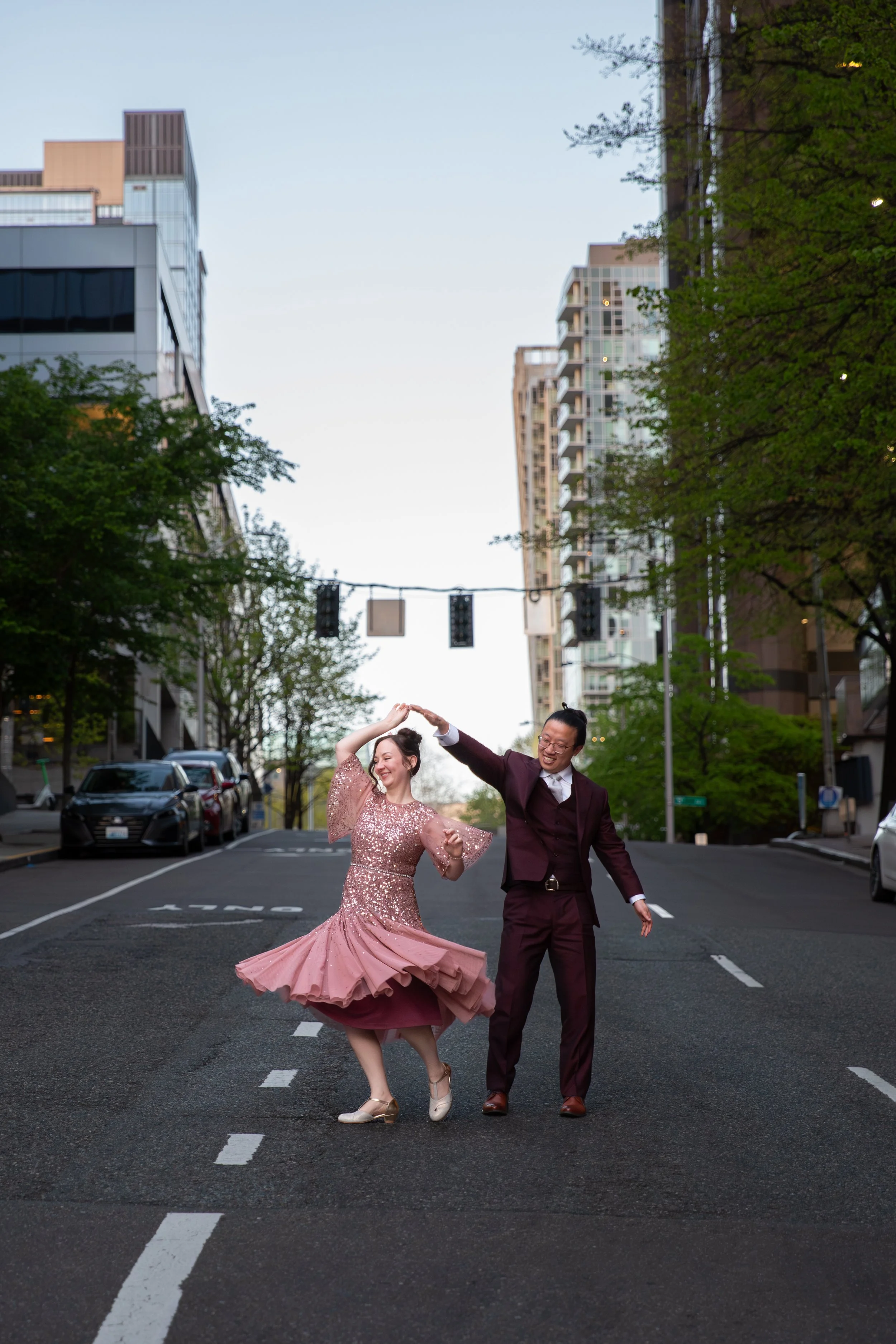 A wedding couple dancing in the middle of a city street during the evening, with tall buildings and green trees in the background. The bride is wearing a pink, sequined dress and the groom is in a dark burgundy suit.