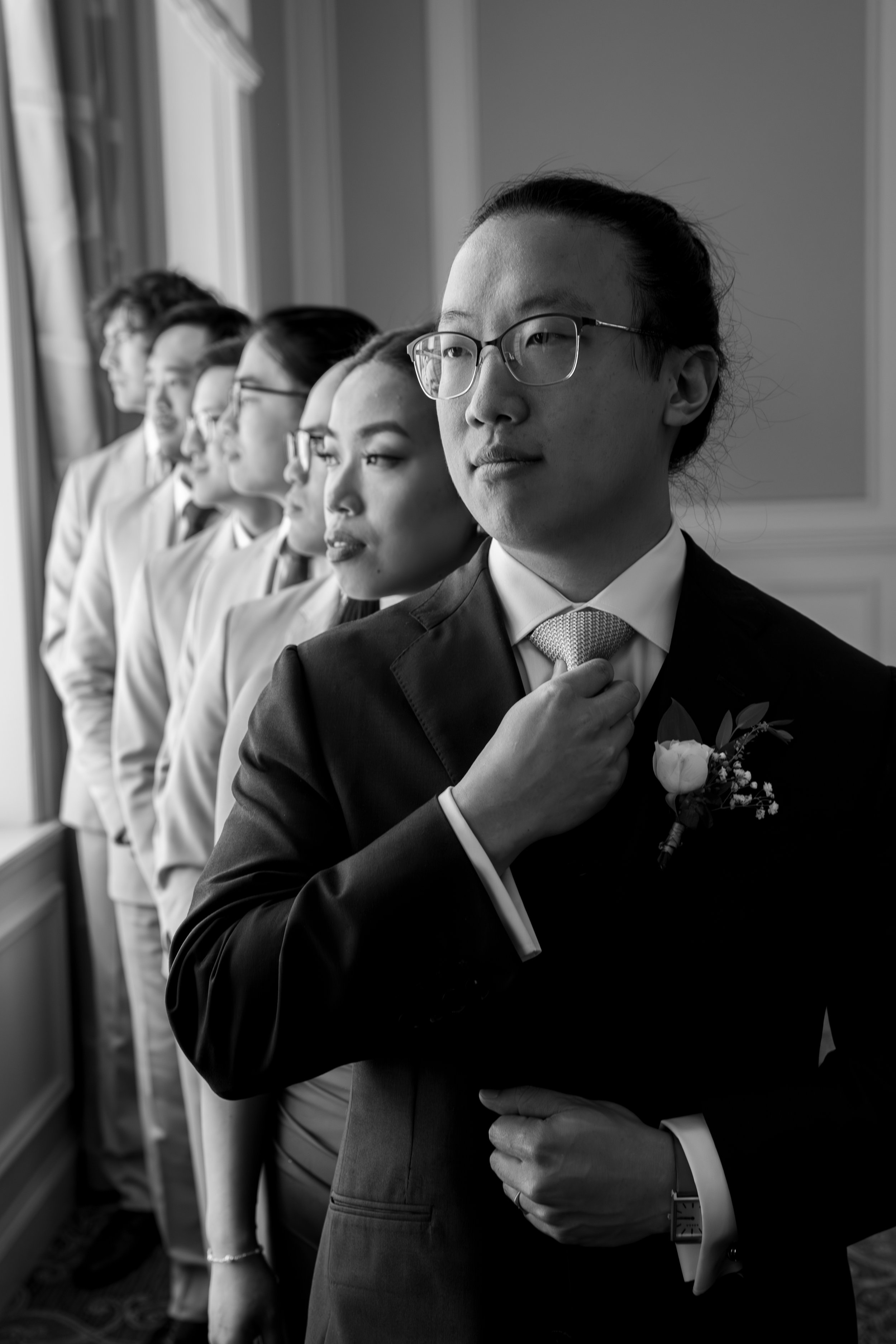 A group of six people in a grooms party with the groom at the front standing in a line by a window, wearing formal attire, with the groom adjusting his suit jacket, during a wedding.