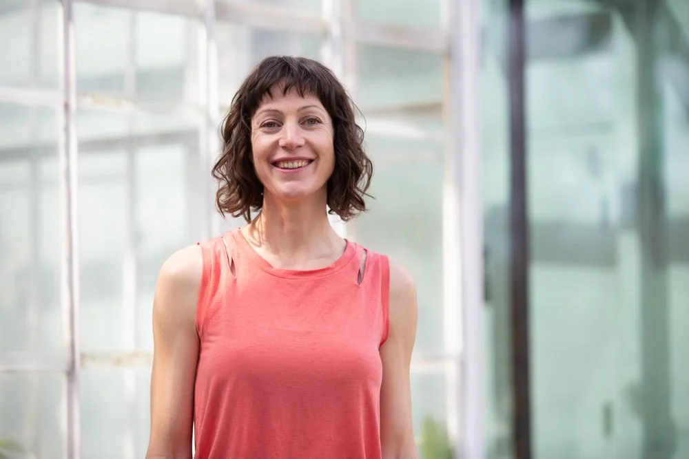 A woman with short brown hair and a pink sleeveless top, smiling, standing in front of a modern glass building during a personal branding photoshoot in Lisbon.