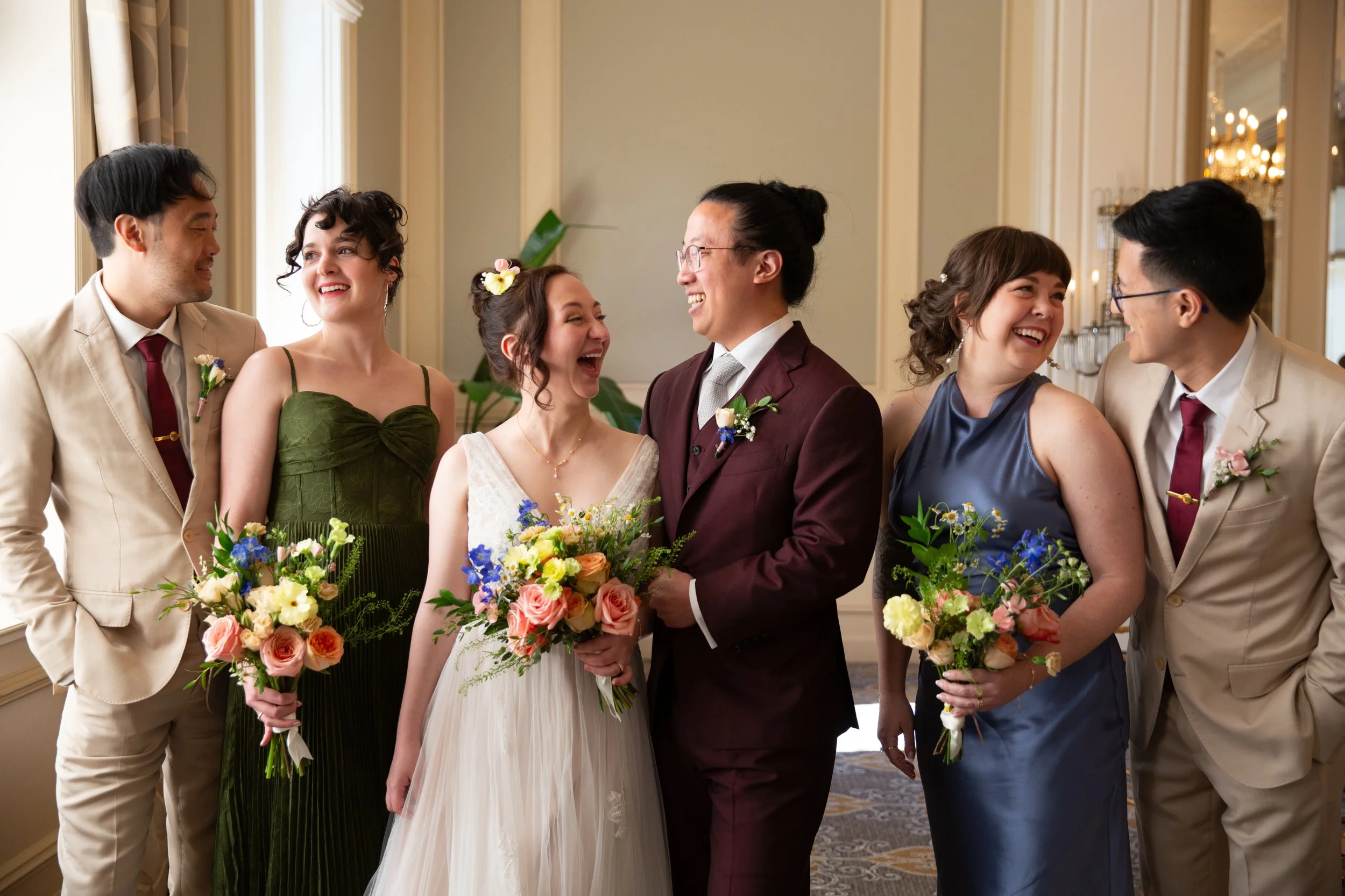 A bride, groom, and four people from their wedding party, dressed in formal attire, holding bouquets of colorful flowers, smiling and engaging with each other in a well-decorated room with large windows and chandeliers at a wedding in Lisbon.