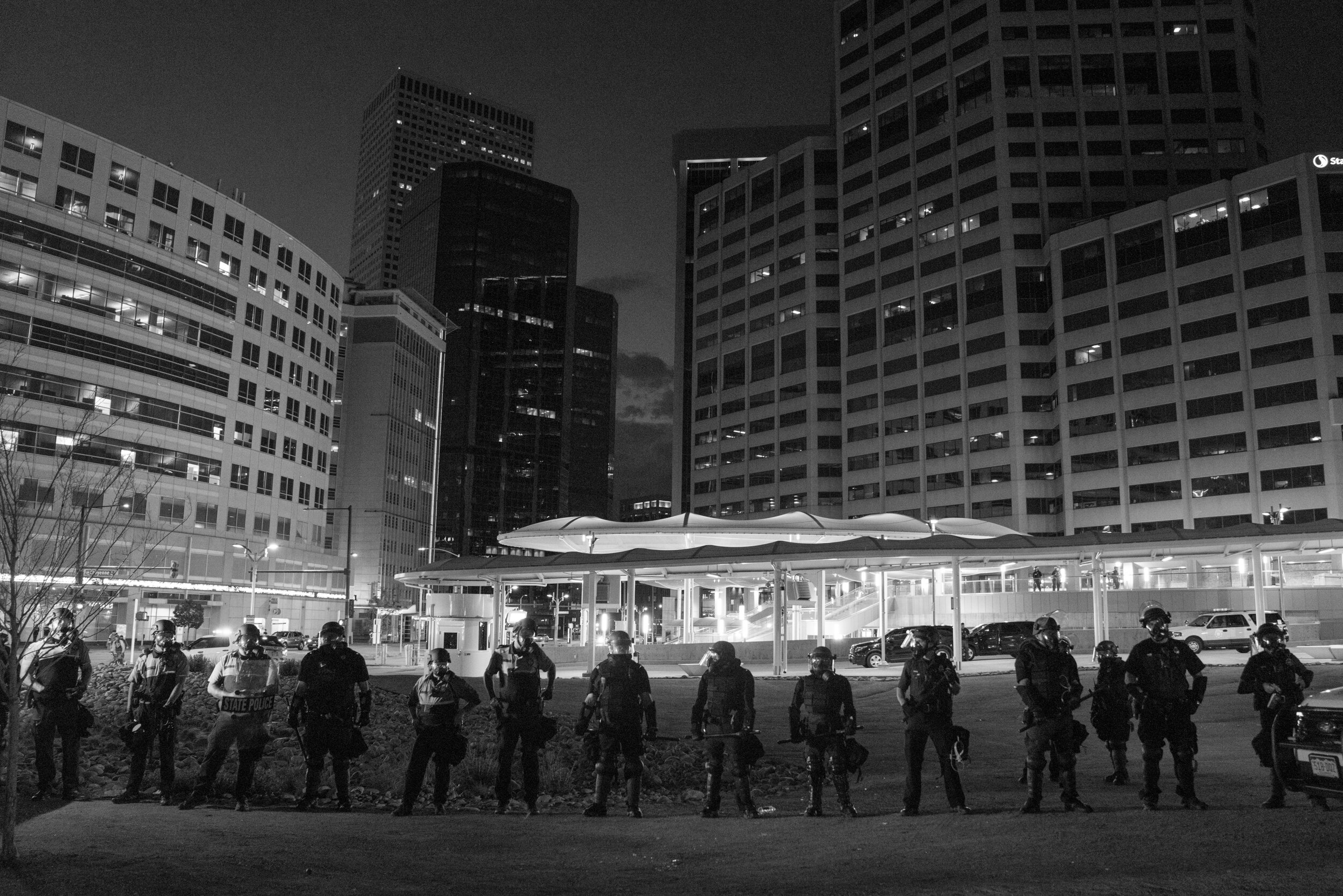Police in riot gear stand in a line with the civic center station and downtown Denver in the background. 5/29/2020.