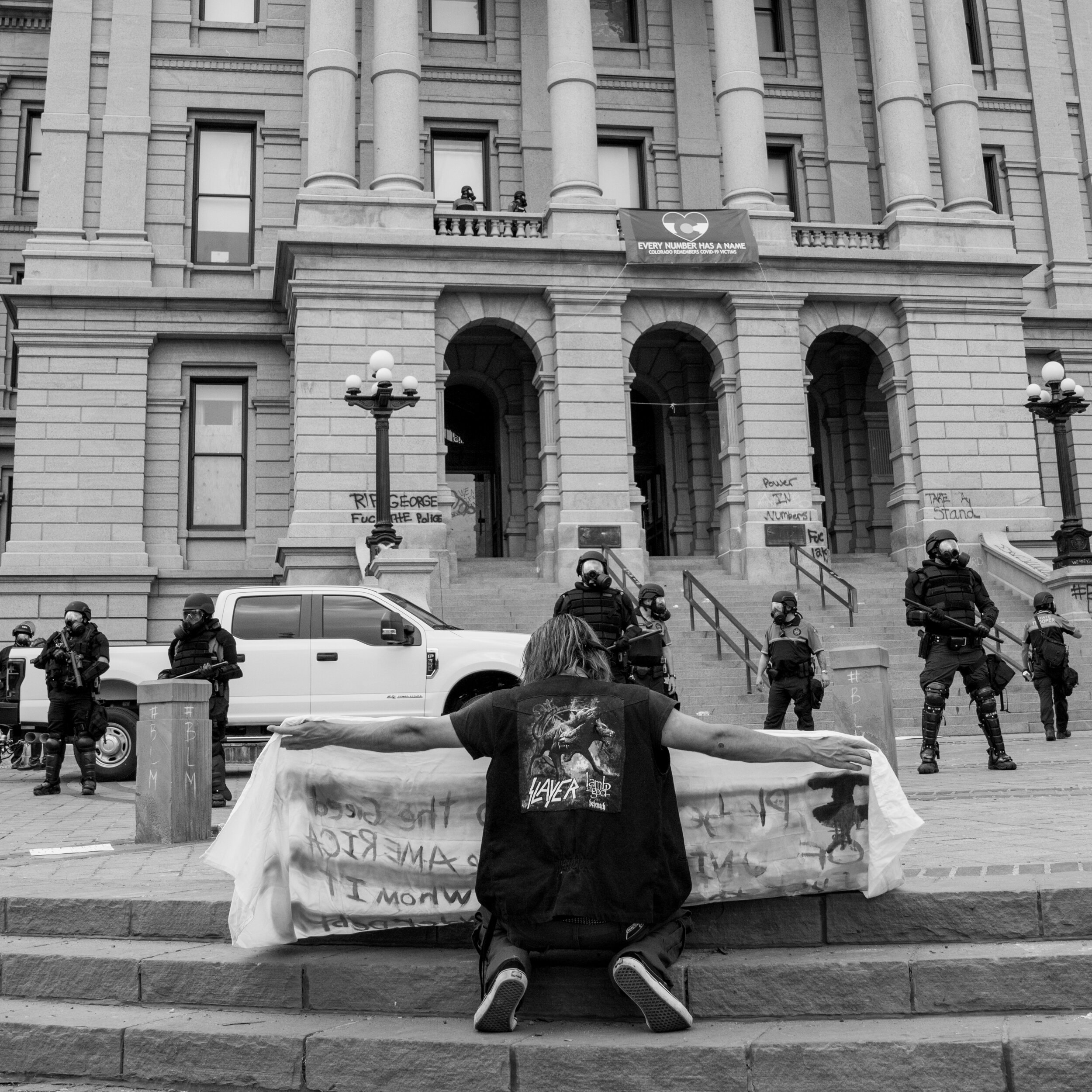 A protester on his knees holds out a sign in front of the vandalized Colorado state capitol building. 5/30/2020.