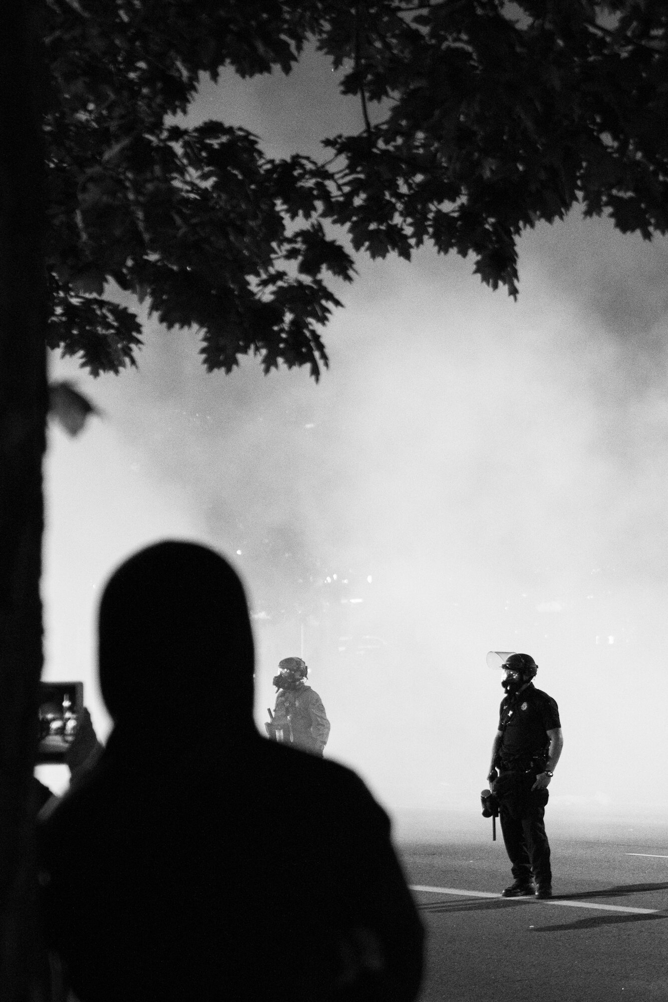 A protester leans against a tree and observes the police while smoke fills the air. 5/29/2020.