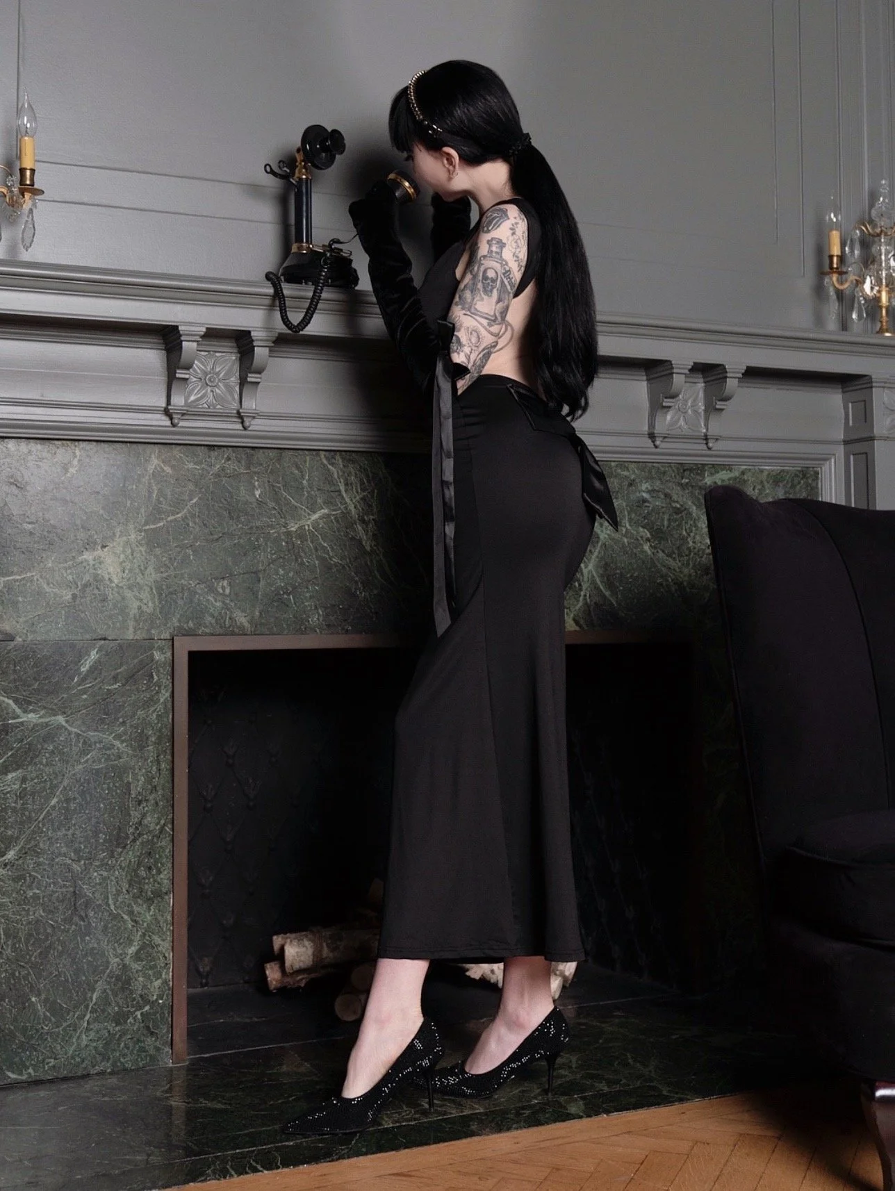 Ramona Ryder NYC Goth Escort is wearing a blakc dress with her hair pulled back facing a wall.