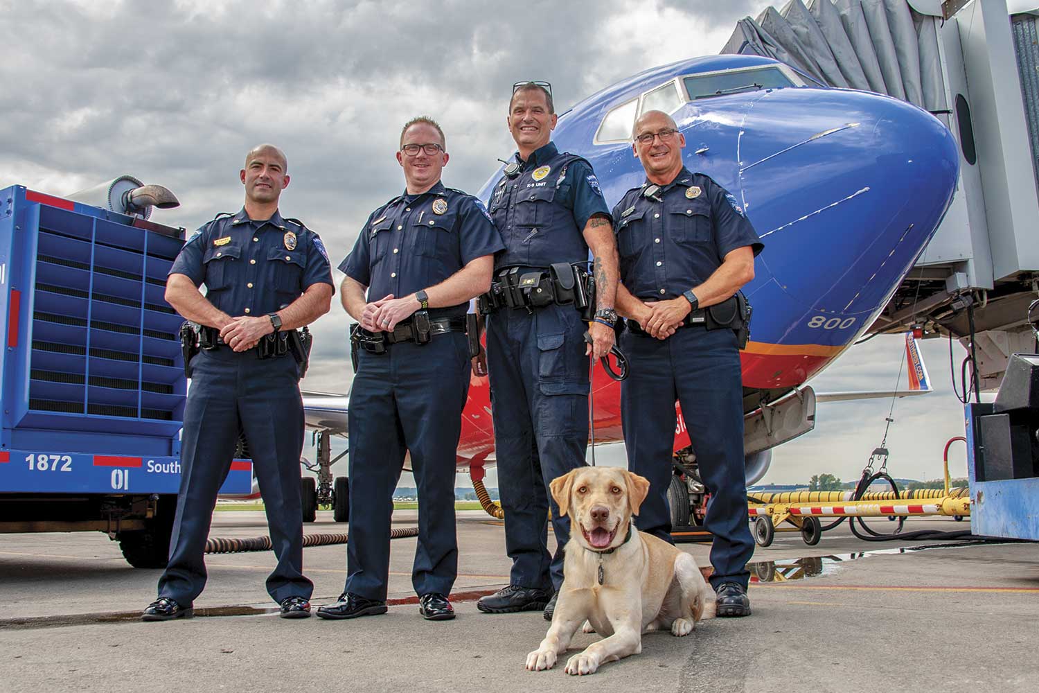  From left-to-right, Maj. Dustin Flannery, Chief Josh Ball, Officer Peter Lamb, K9 Raj and Officer Jeff Rogers pose in front of a Southwest Airlines plane on the tarmac at the Louisville International Airport. (Photo by Jim Robertson) 
