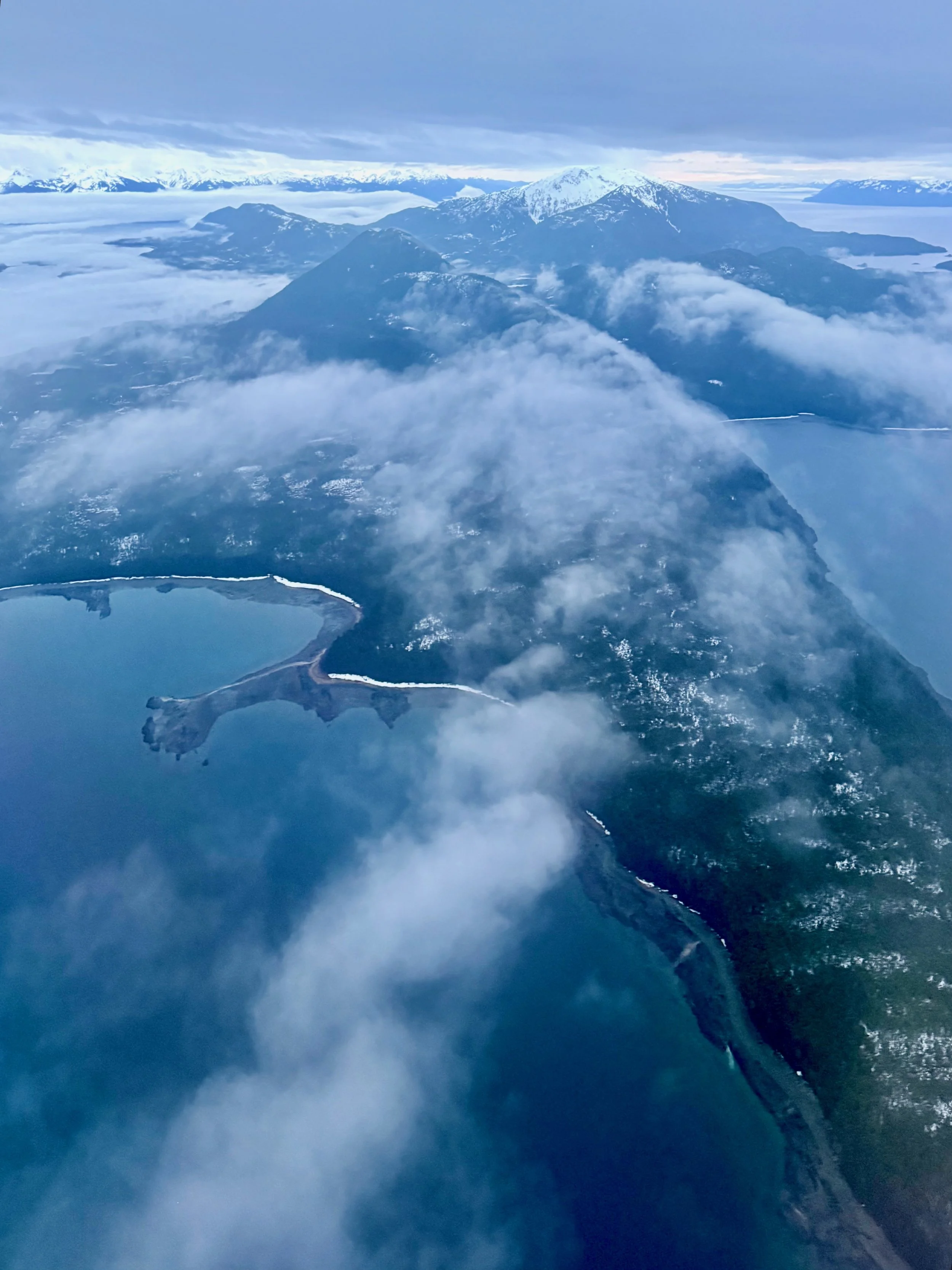 Aerial view of snow-capped mountains, clouds, and a body of water with a peninsula or island in the foreground.
