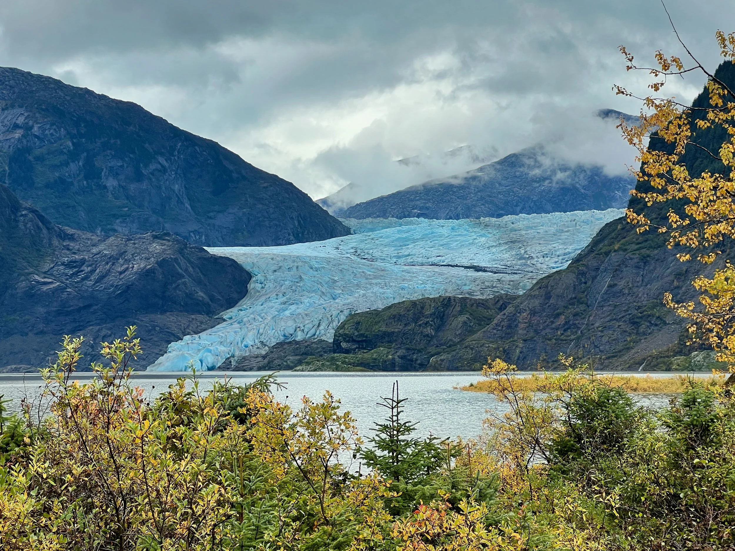 A scenic mountain landscape featuring a glacier, with lush greenery in the foreground, mountain peaks partly covered by clouds, and a body of water at the base.