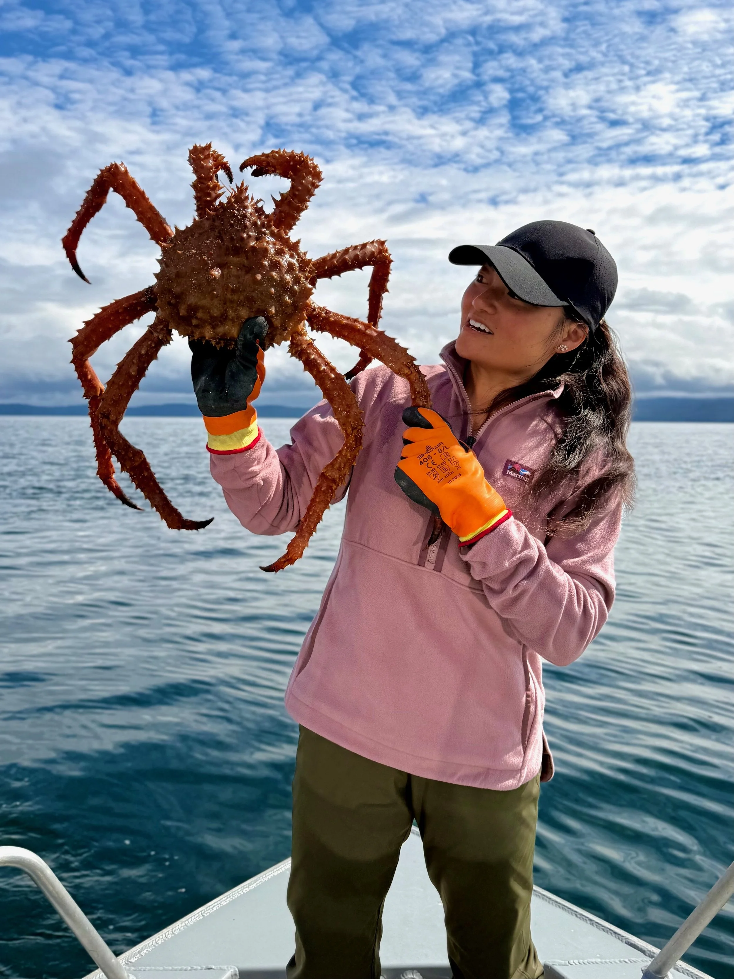 A woman wearing a pink jacket and orange gloves holding a large crab on a boat in a body of water with a cloudy sky in the background.