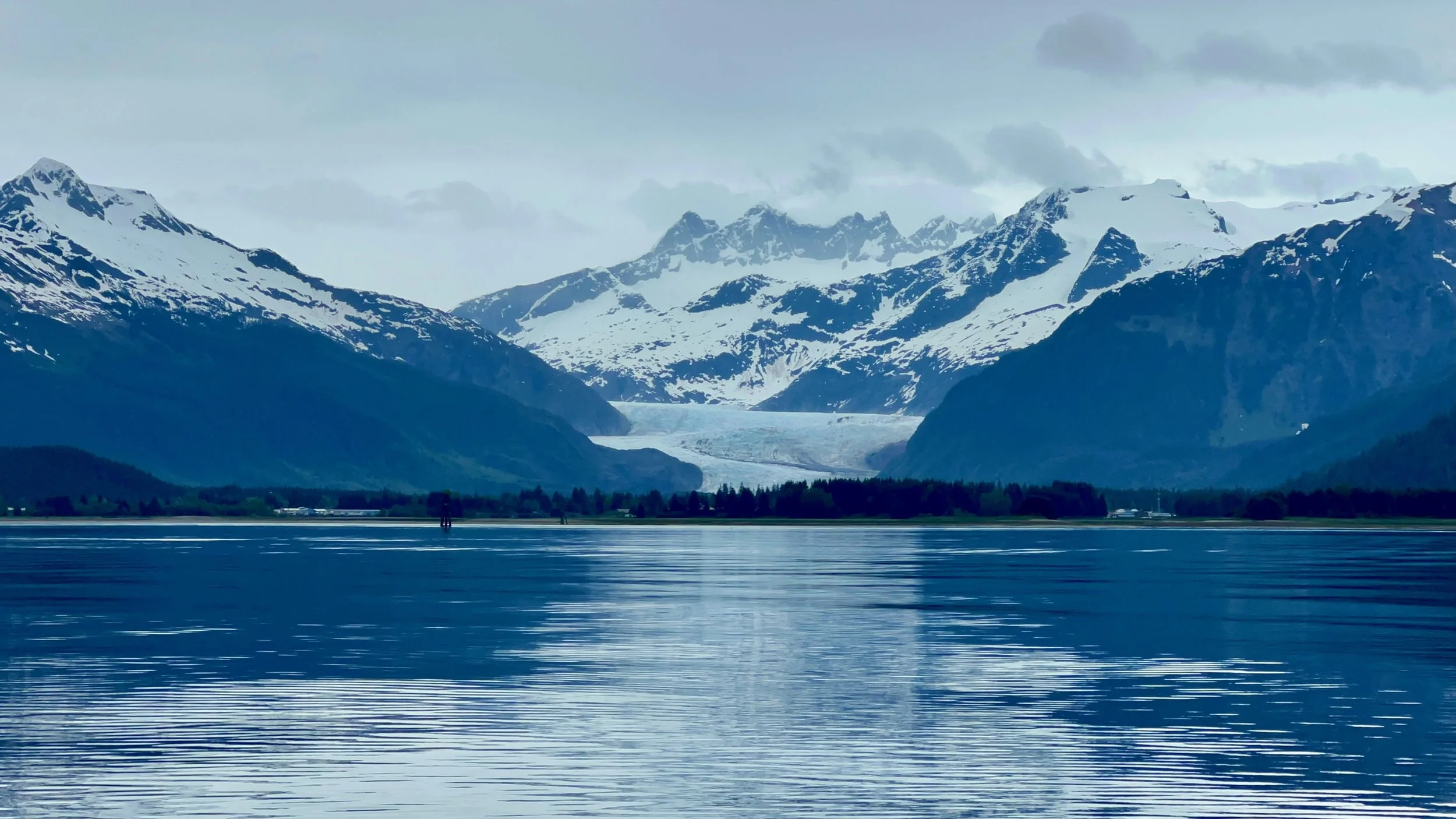Snow-capped mountains behind a lake with a small group of people on the shore in the foreground.