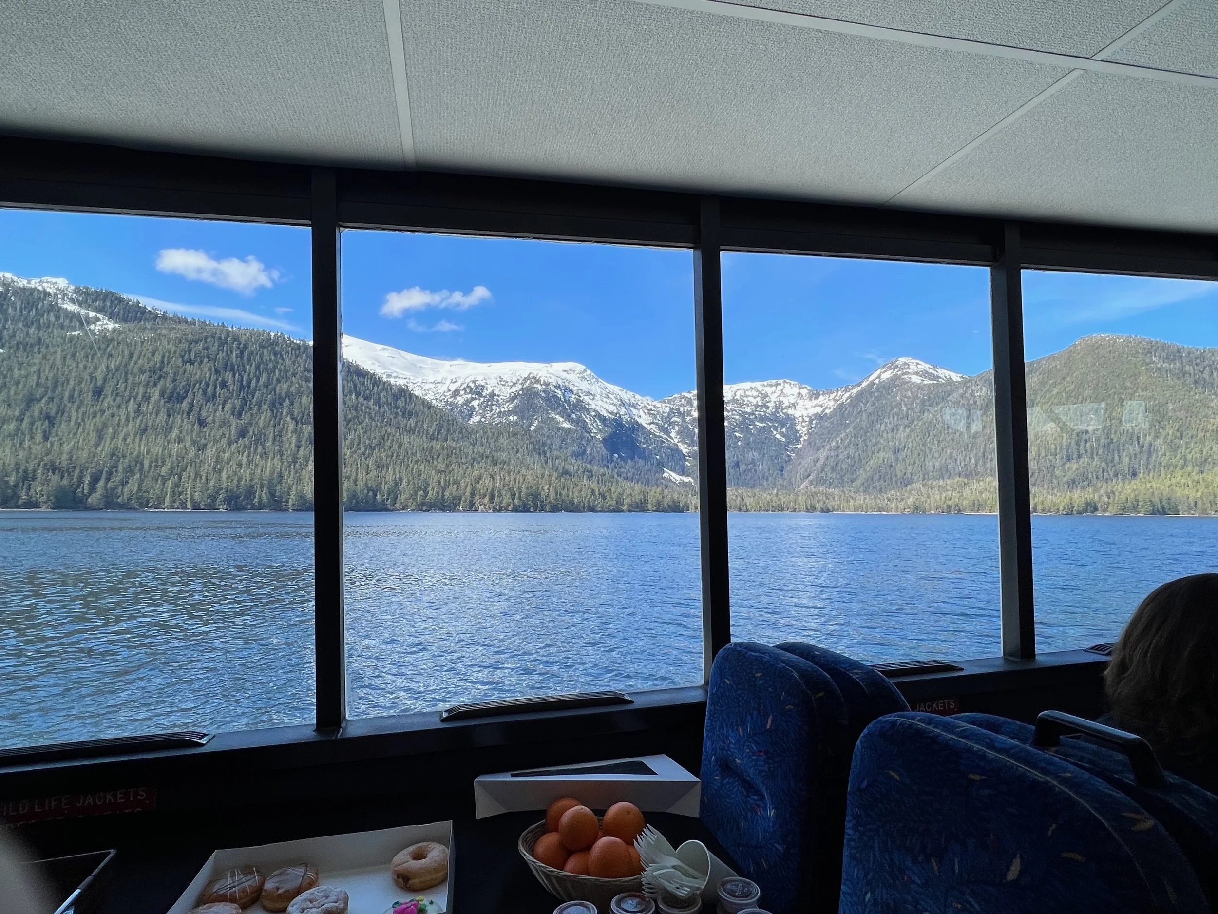 View of a mountain lake through a window in a boat or ferry with snow-capped mountains and a partly cloudy blue sky, and some snacks and orange in the foreground.