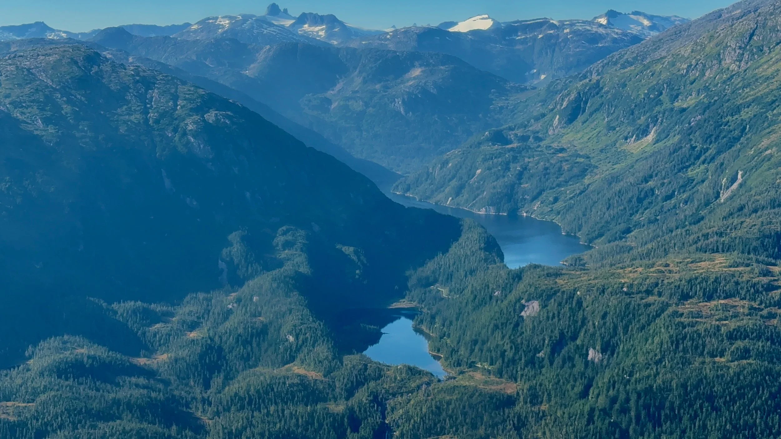 A view of a large mountainous landscape with snow-capped peaks in the distance, green forested valleys, and a winding river or lake running through the valleys.