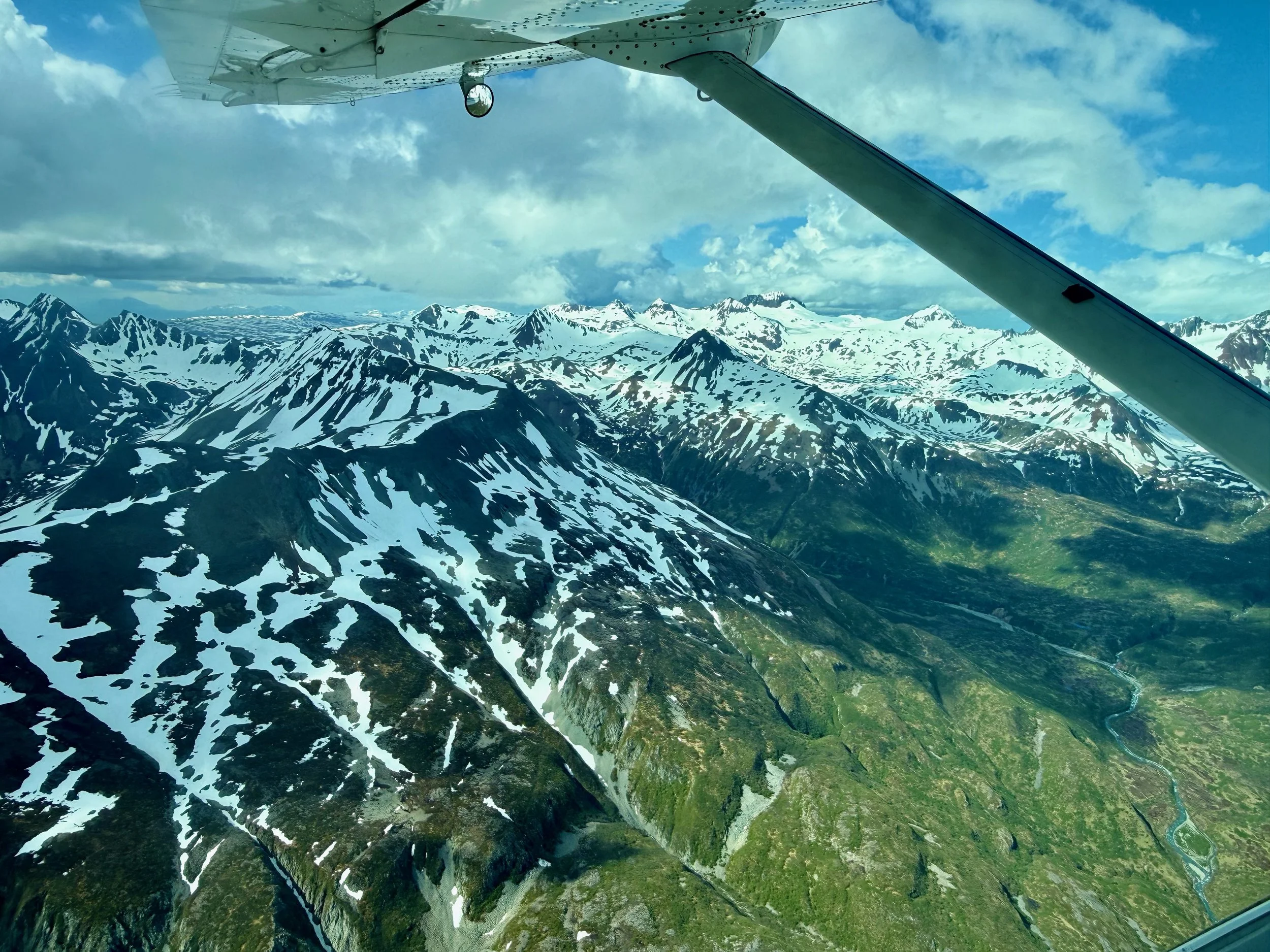 A view from an airplane window shows snow-capped mountains under a partly cloudy blue sky.