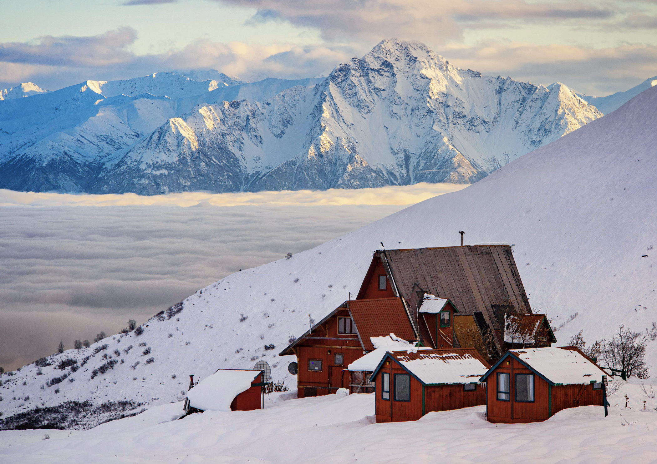 Snow-covered mountain landscape with a cluster of wooden houses in the foreground, isolated amid snow and fog, with a backdrop of rugged snow-capped peaks and a partly cloudy sky.