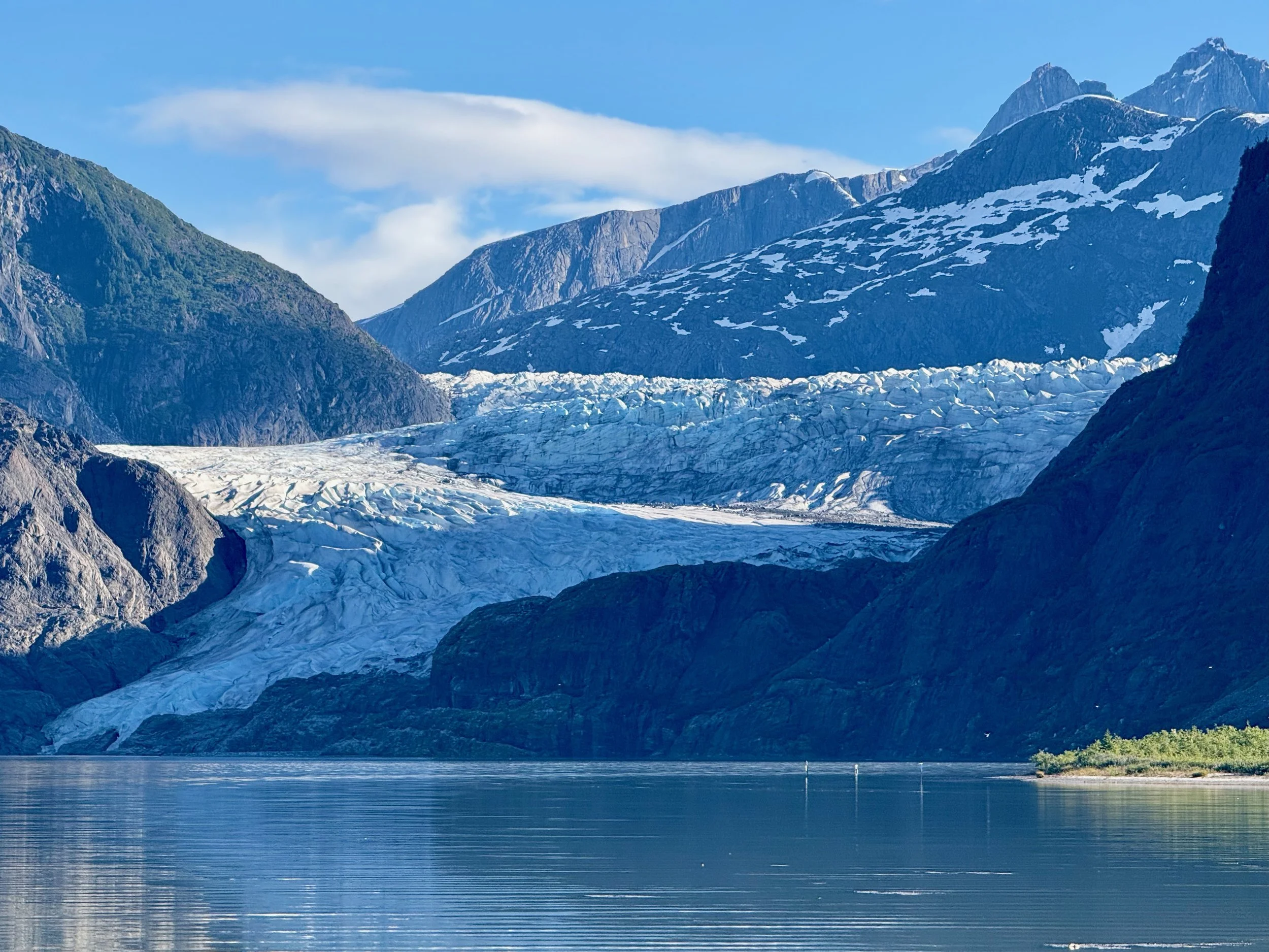 A scenic landscape featuring snow-capped mountains, a glacier, and a calm body of water in the foreground, with a blue sky and some clouds.