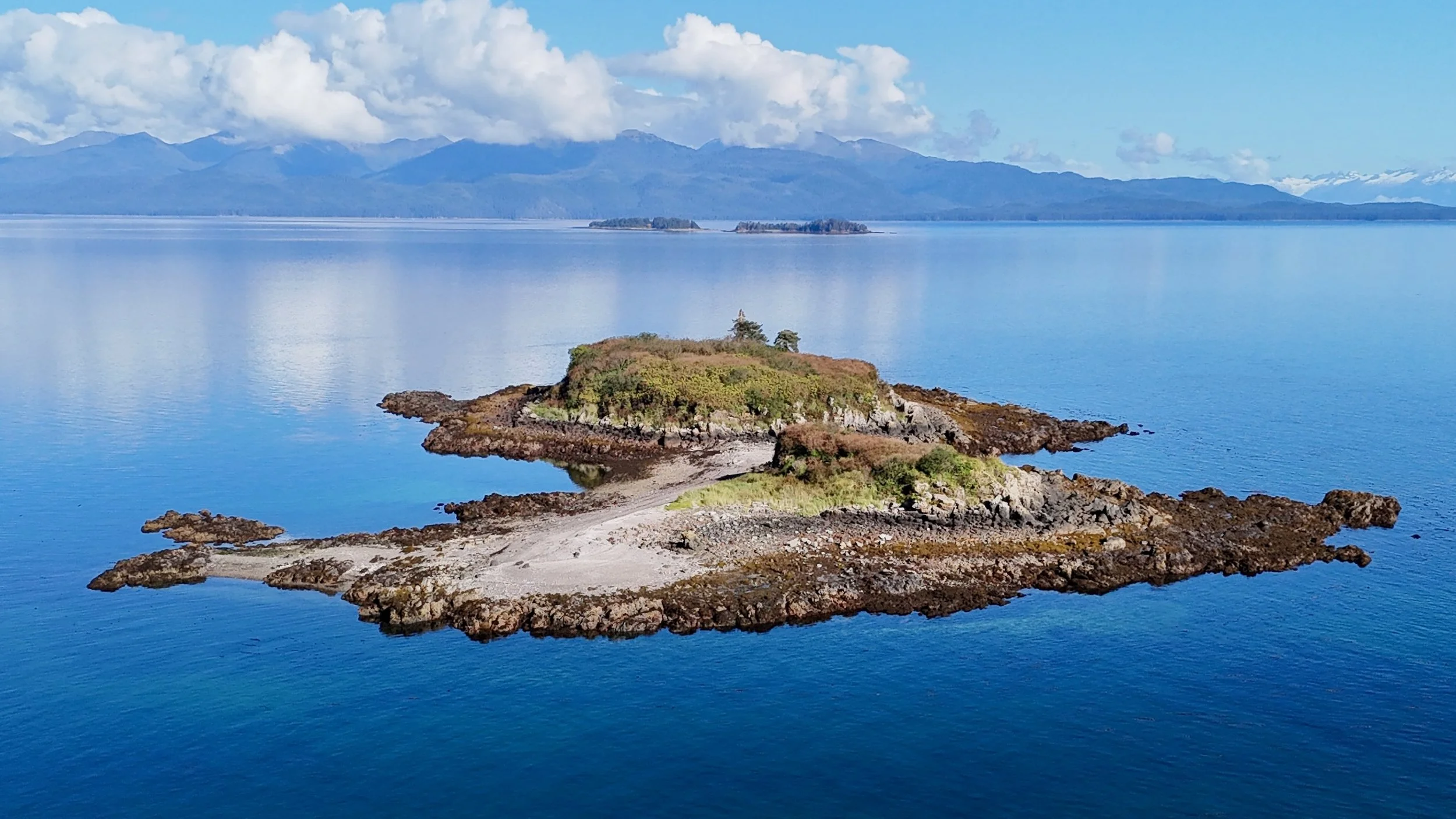 A small island with rocky shores and greenery in a calm blue body of water, with mountains and clouds in the background.