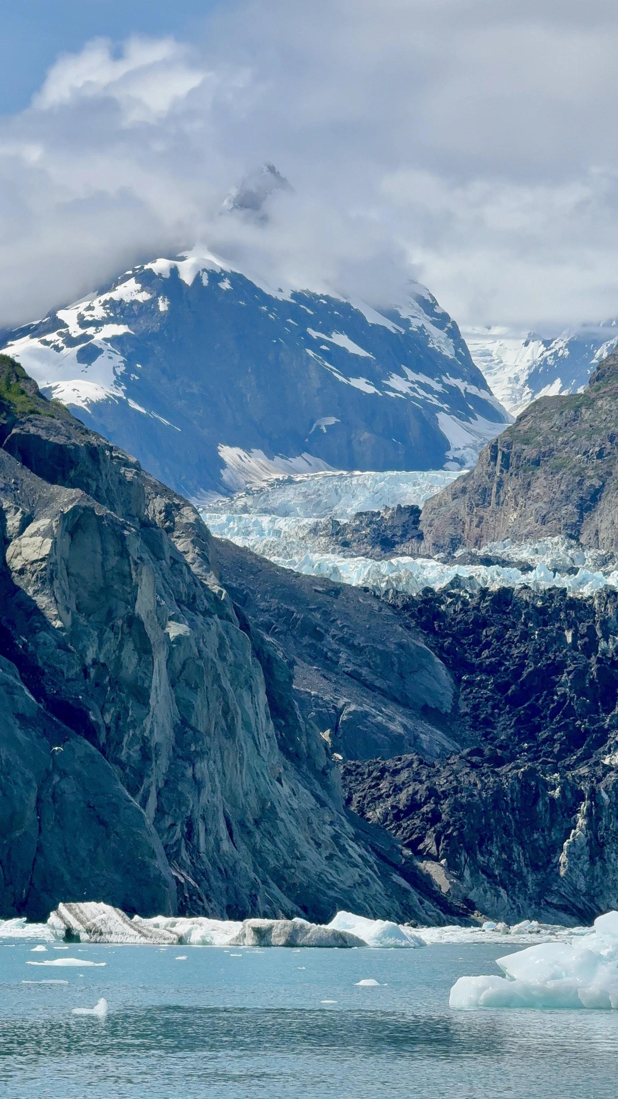 Snow-capped mountains partially covered in clouds, glacier formations, and floating ice in a calm icy body of water.