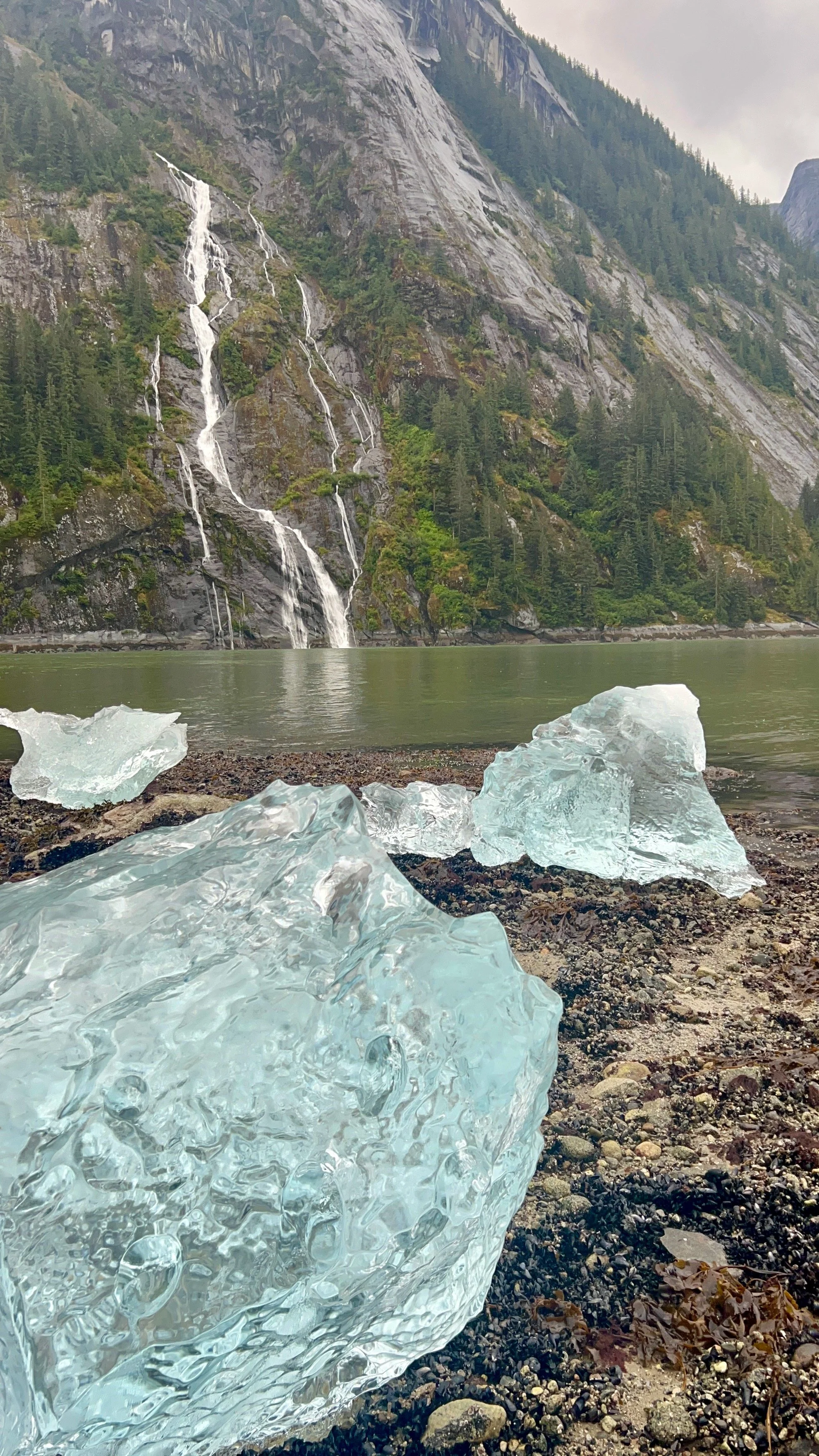A landscape scene with a mountain, multiple waterfalls, a lake, and large ice chunks on a rocky shore.