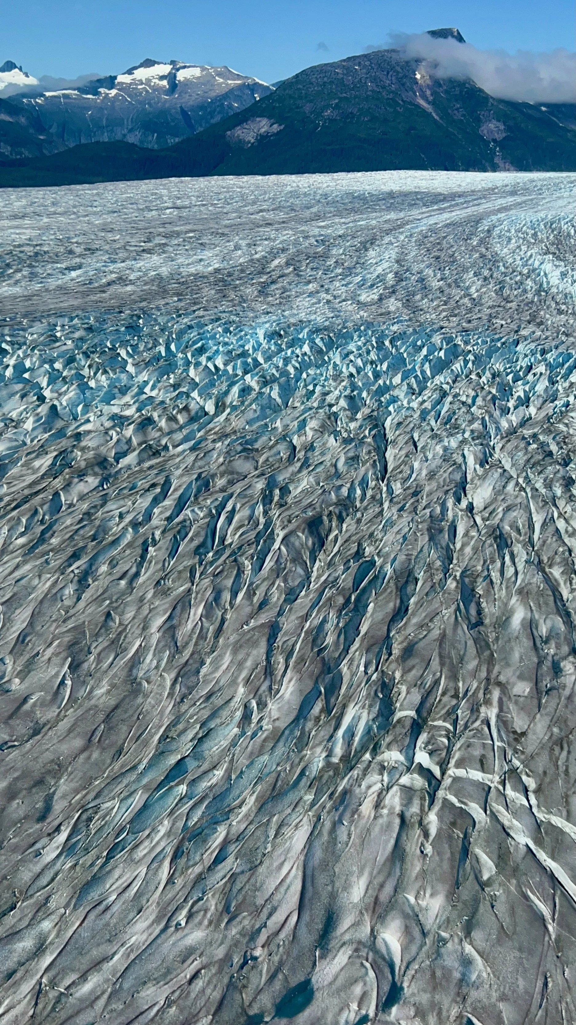 A vast glacier with deep blue crevasses leading towards snow-capped mountains in the background under a clear blue sky.