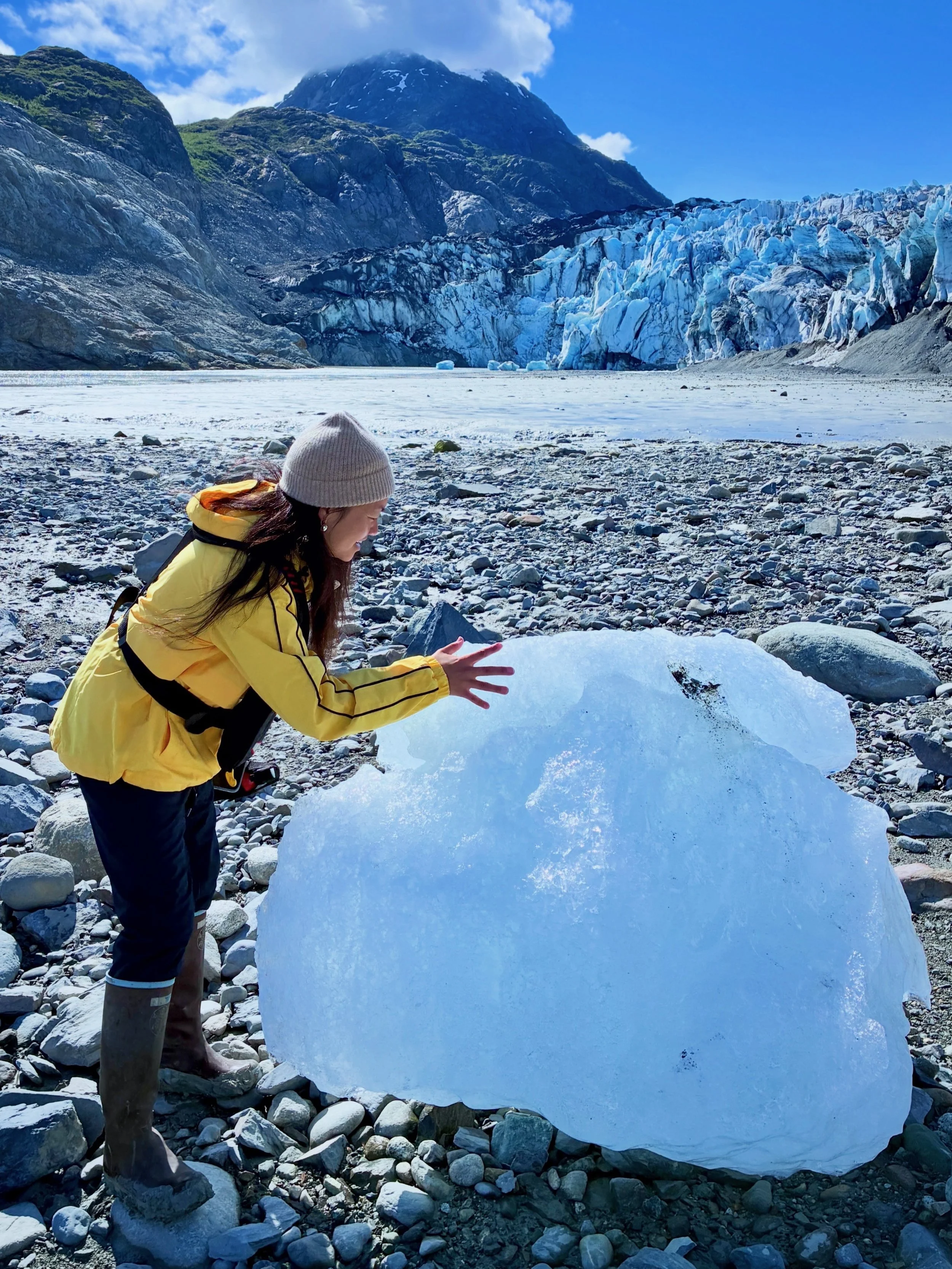 Mary Jacquel wearing a yellow jacket, gray beanie, and boots is kneeling on rocky terrain, touching a large piece of ice. In the background, there is a glacier with blue ice and surrounding mountains under a clear blue sky with a few clouds.