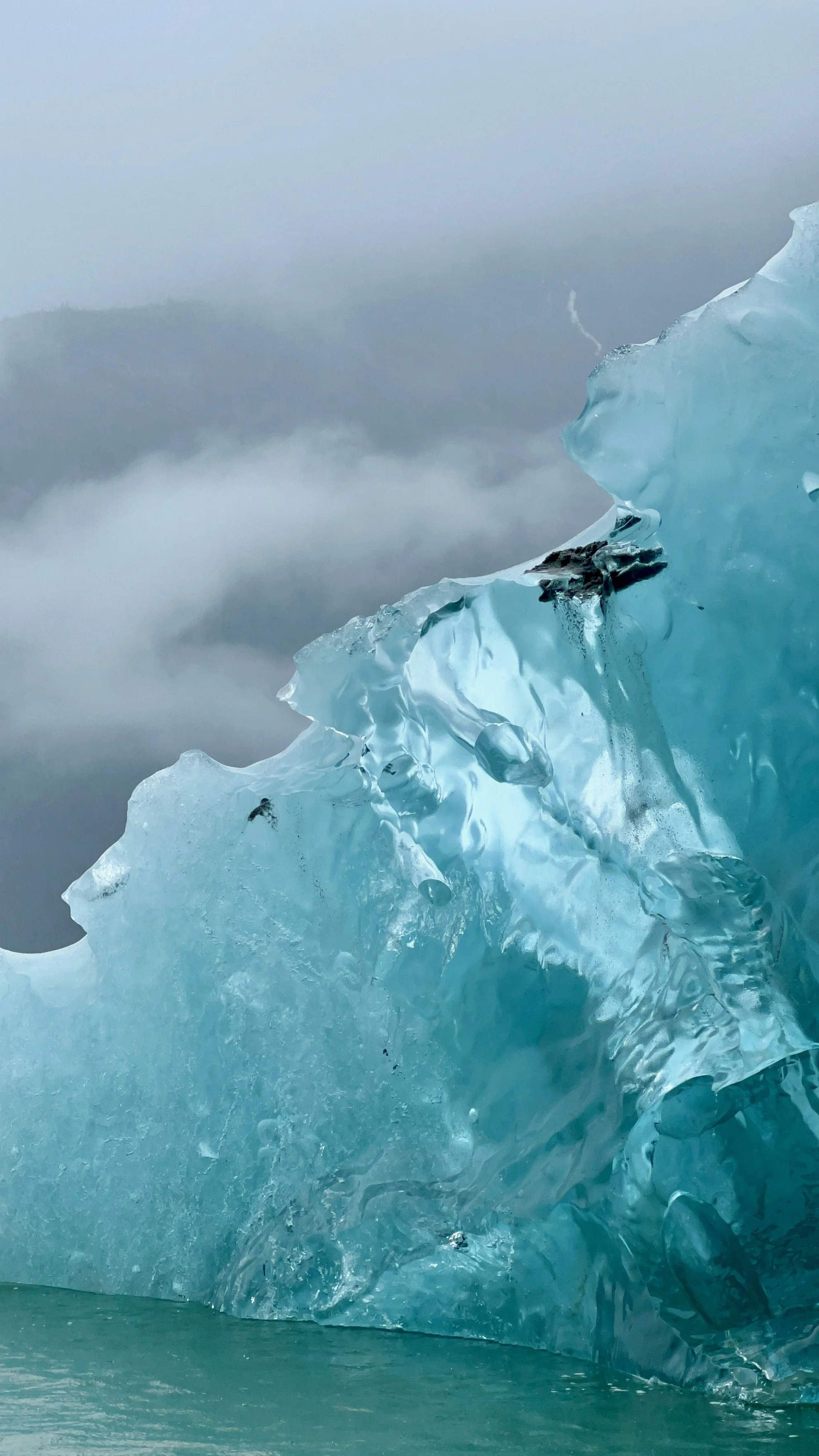 A large blue glacier with a rough and jagged surface, set against a foggy overcast sky in icy terrain.