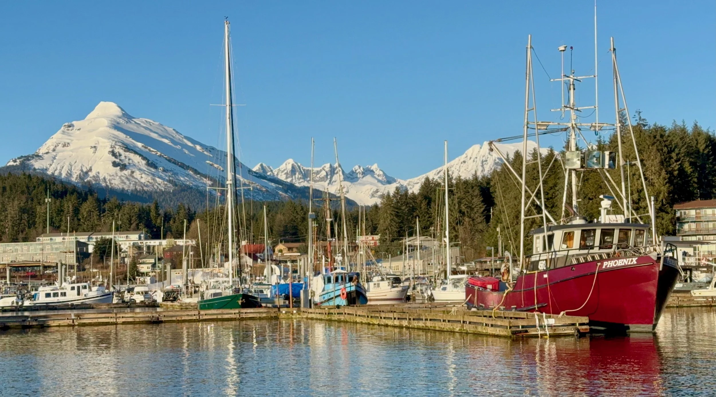 A marina with several boats docked, including a red boat named 'Phoenix,' with snow-capped mountains and a town in the background under a clear blue sky.