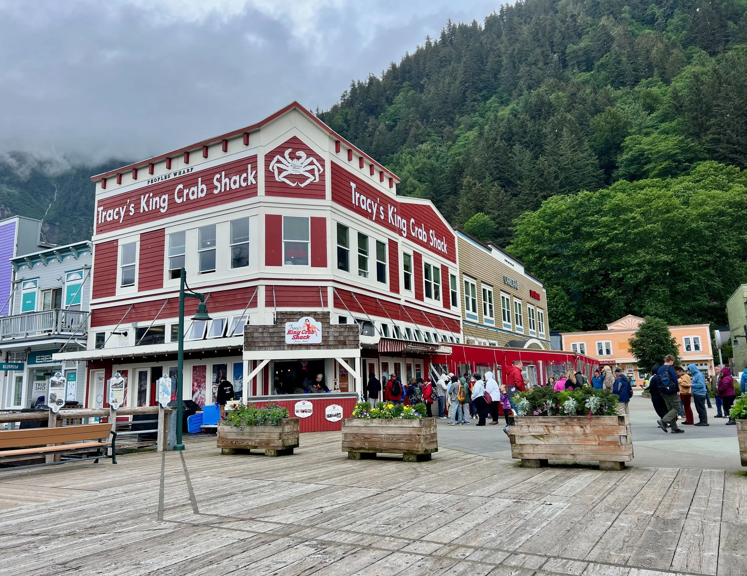 A colorful waterfront building with signs for Tracy's King Crab Shack, surrounded by people, benches, and flower planters, with a lush green mountain in the background.