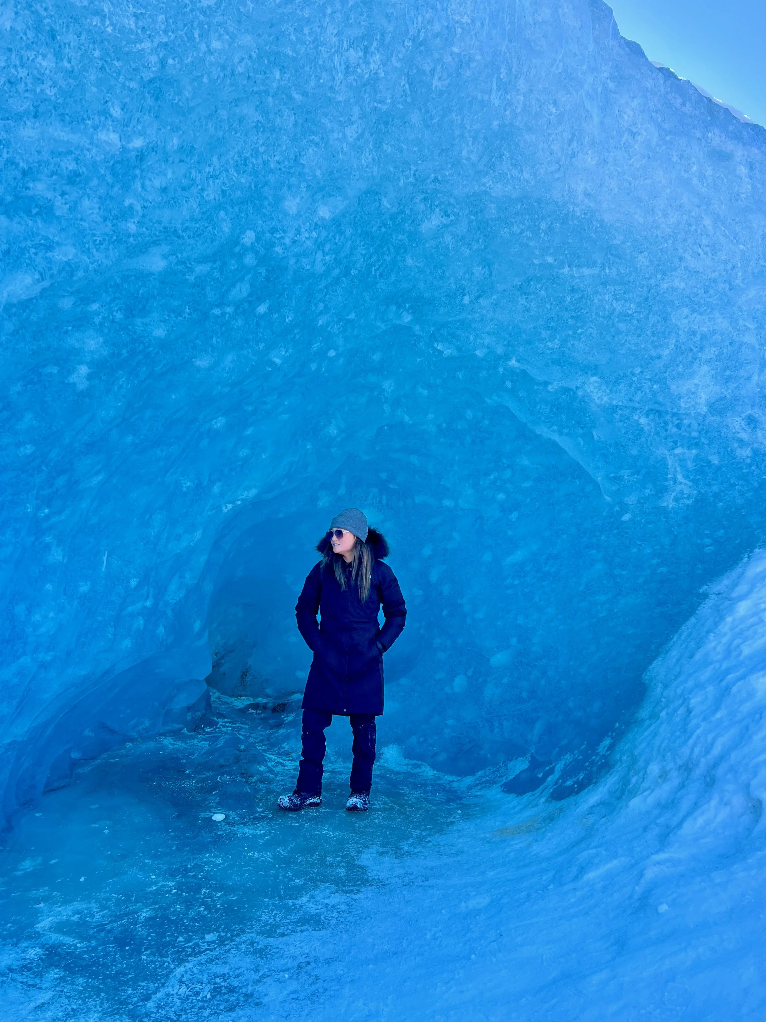 A woman standing inside an ice cave, wearing a dark coat, grey beanie, sunglasses, and boots, surrounded by ice walls and ceiling