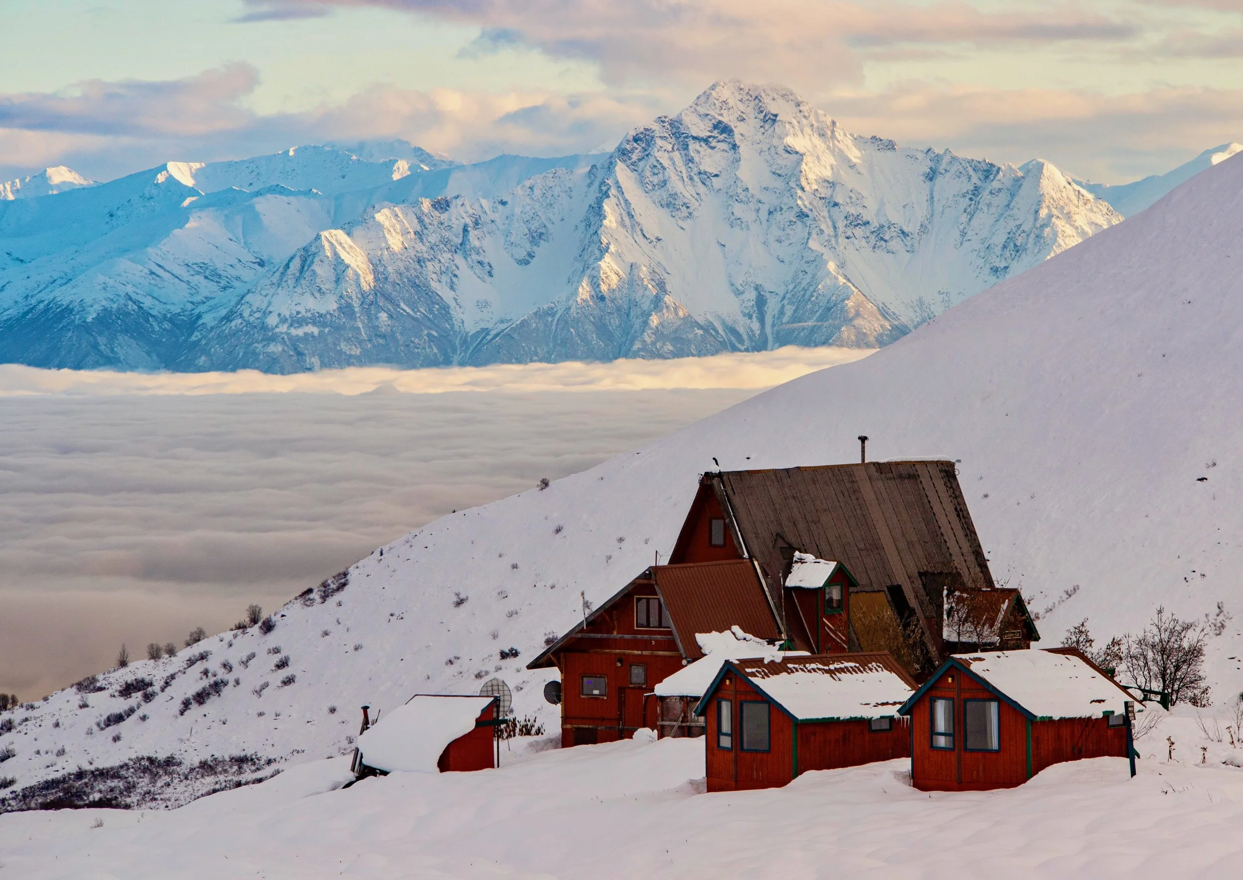 Snow-covered houses on a hillside with a mountain range in the background under a partly cloudy sky.