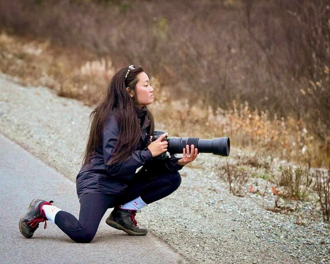 Mary Jacquel kneeling on roadside, holding a camera with a large telephoto lens in Denali National Park, in an outdoor setting with dry foliage in the background.