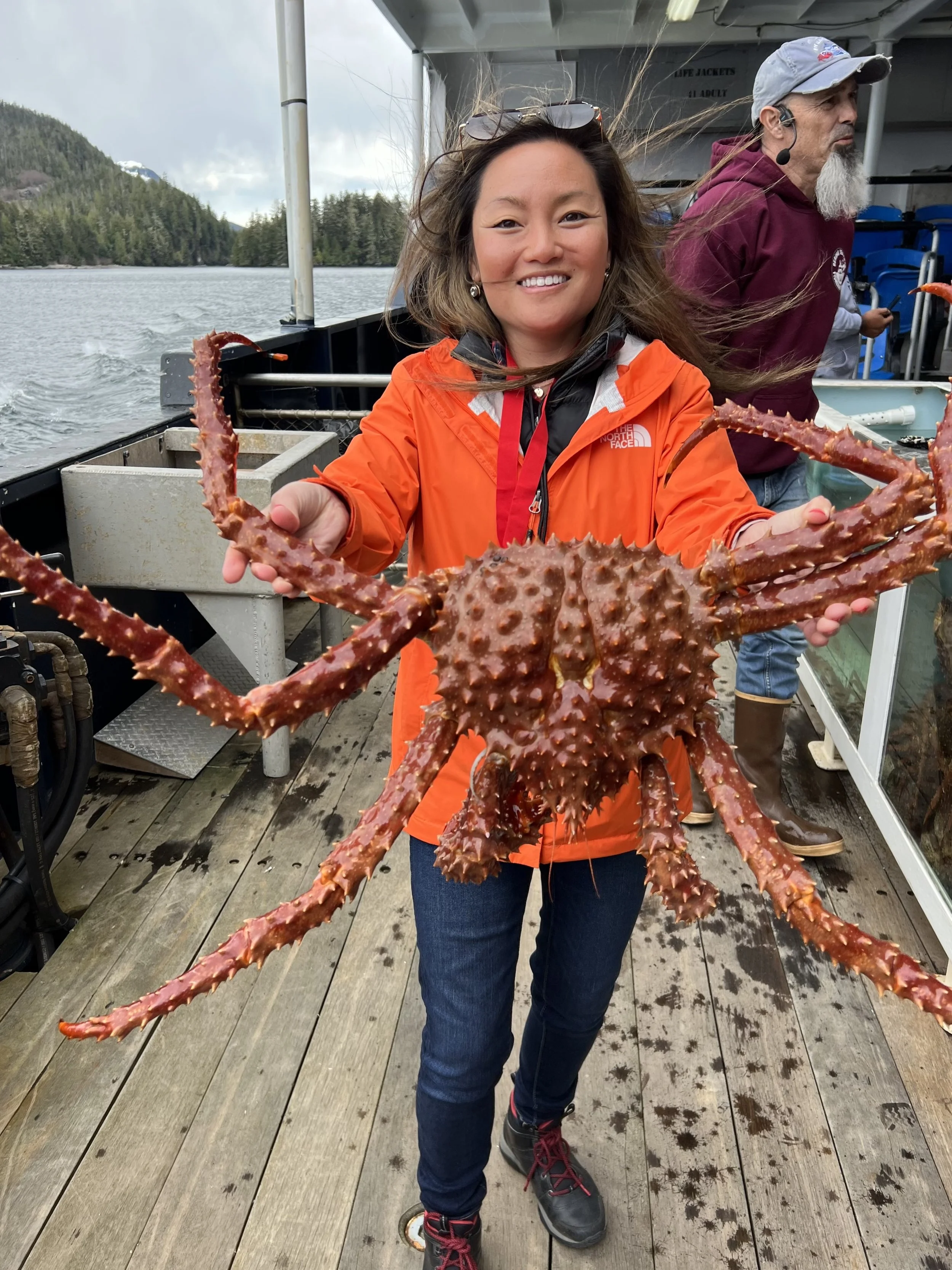Woman in orange jacket holding large king crab on boat with waterfront and trees in background.