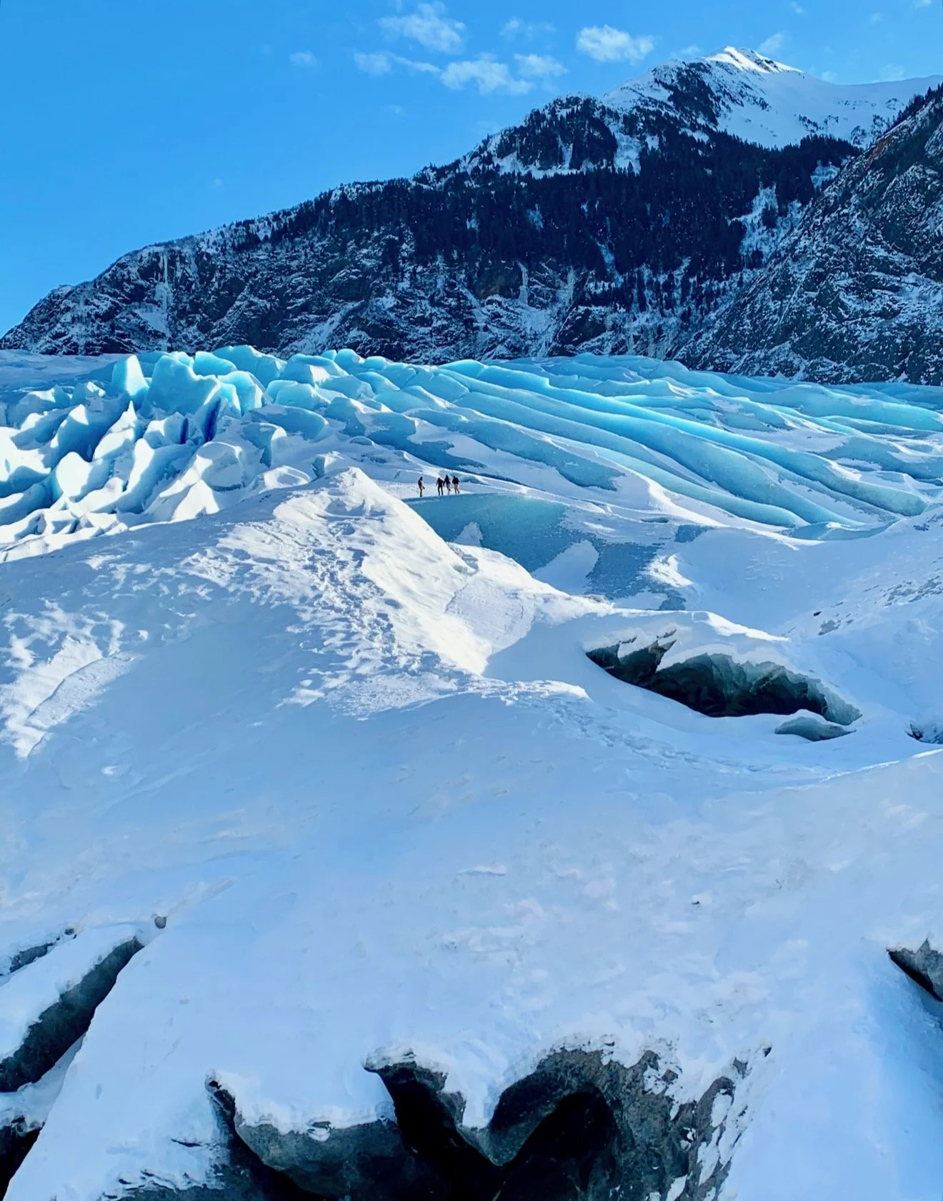 Three hikers walking across a snow-covered glacier, with a mountain range in the background and a snow-capped mountain peak in the distance.