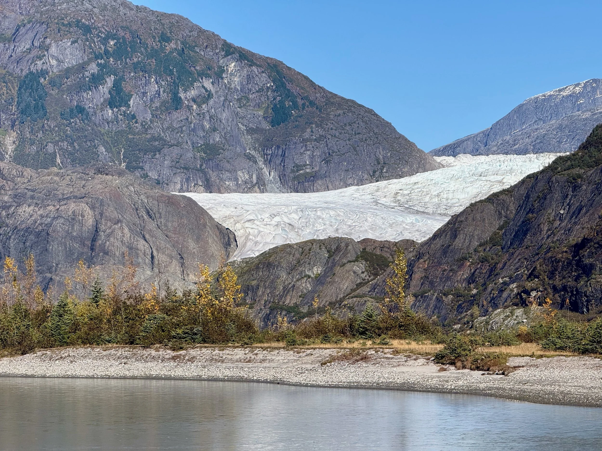 A landscape of mountains with a glacier, green trees, and a body of water in the foreground.