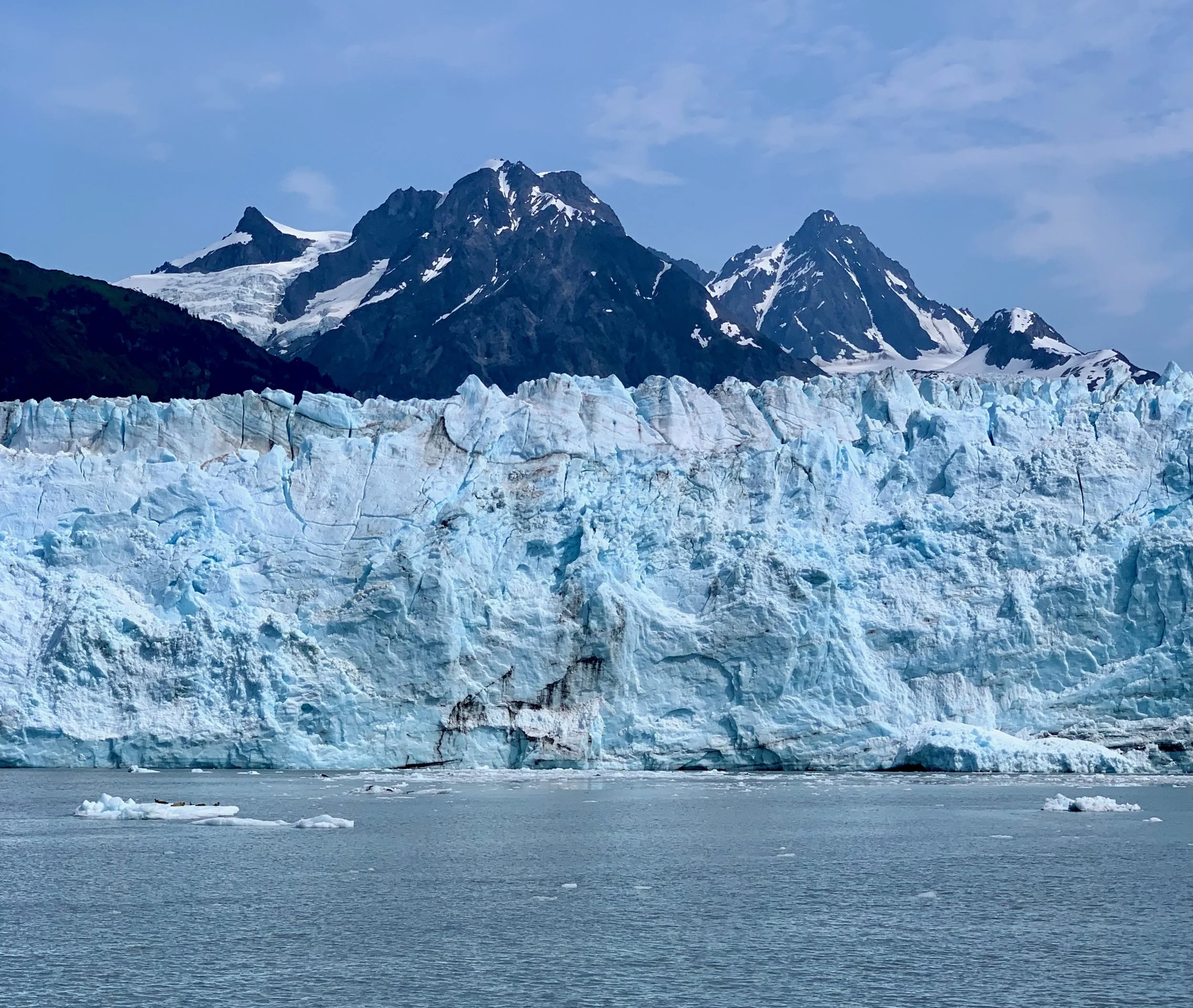 View of a glacier with towering snow-capped mountains and a partly cloudy sky in the background. The glacier has a jagged ice surface and appears to be calving into a body of water with floating ice chunks.
