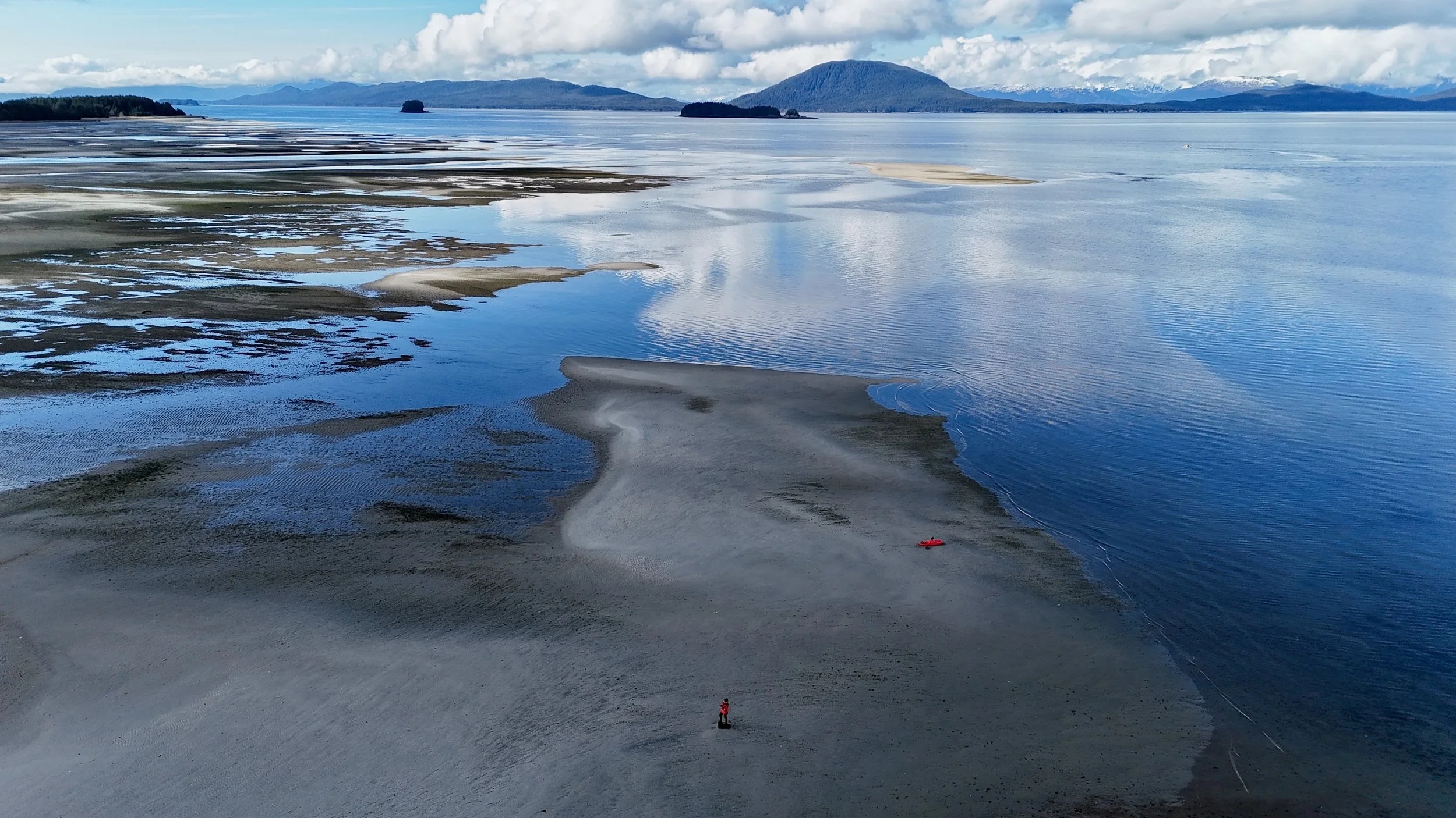 A wide view of a coastal area with sand and shallow water in the foreground. In the distance, there are small islands and mountains under a cloudy sky.