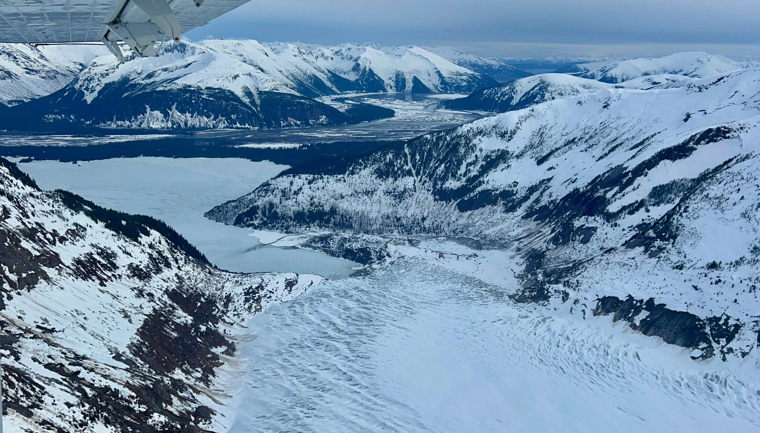 Aerial view of snow-covered mountains and glaciers with a river winding through the valley in the distance, under a cloudy sky.