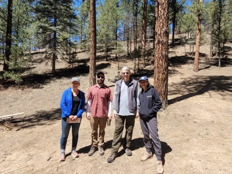 Four individuals in hiking boots and jackets pose for the camera in front of an open-canopy pine forest