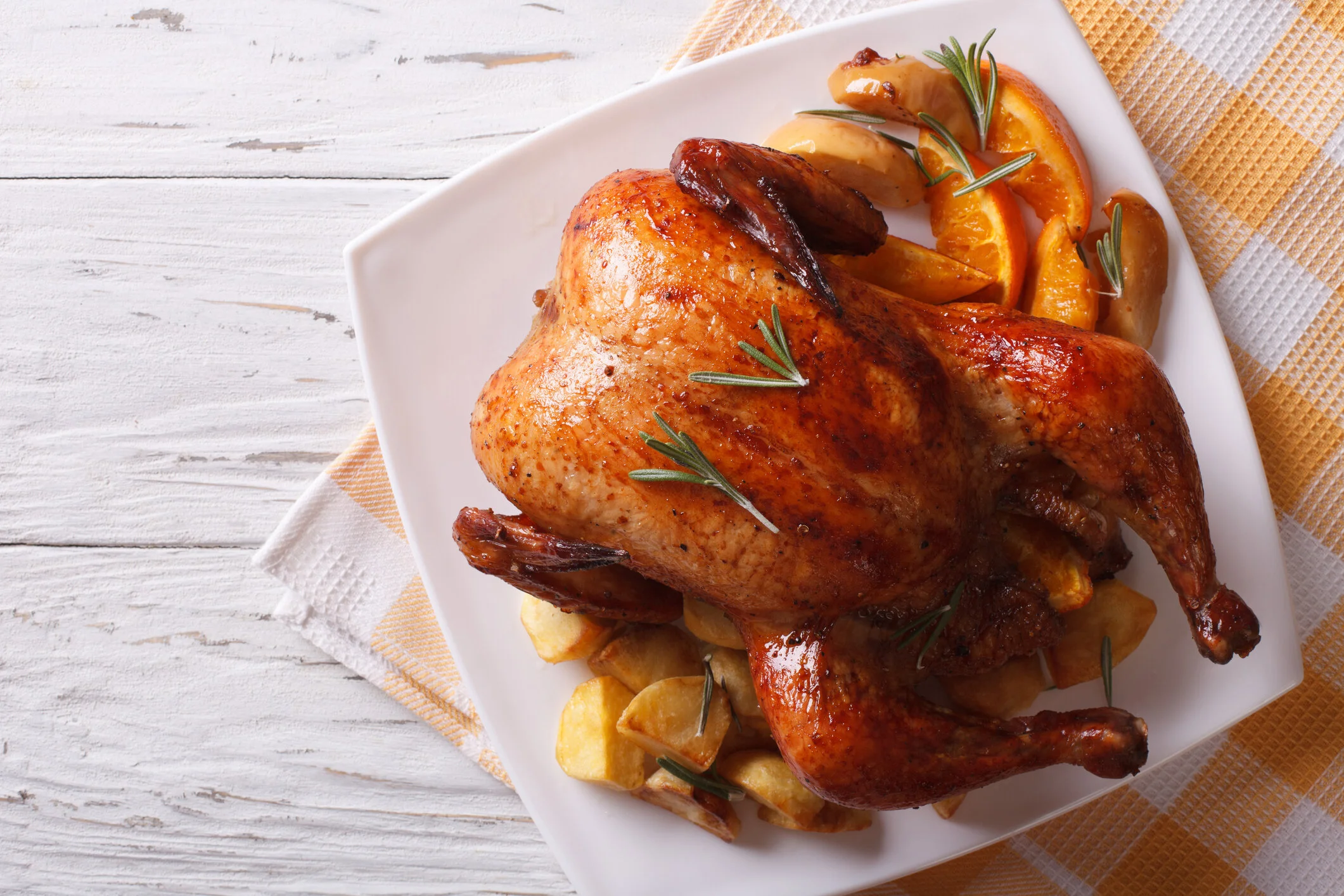 PHOTO DESCRIPTION: Fully roasted turkey with roasted potato and citrus garnish on a square white plate, placed on a checkered tea towel on top of a shiplap shite table.