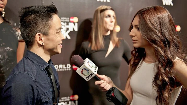 A woman holding a microphone interviews a smiling man at a red carpet event, with a woman in a black dress in the background.