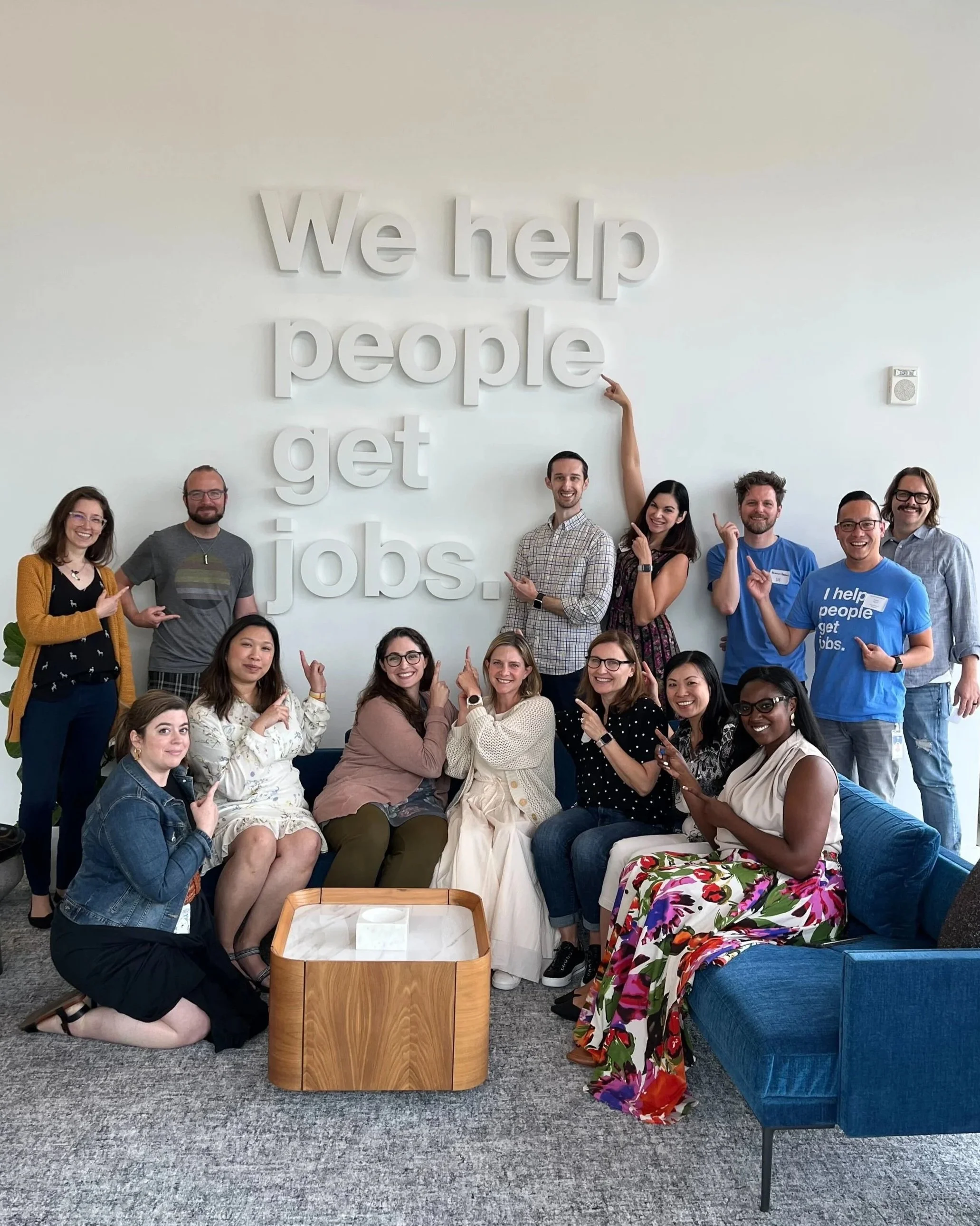 Group of diverse people posing together in front of a wall with white 3D letters that say 'We help people get jobs.' The group is smiling and pointing at the sign, with some standing and others sitting around a wooden table.