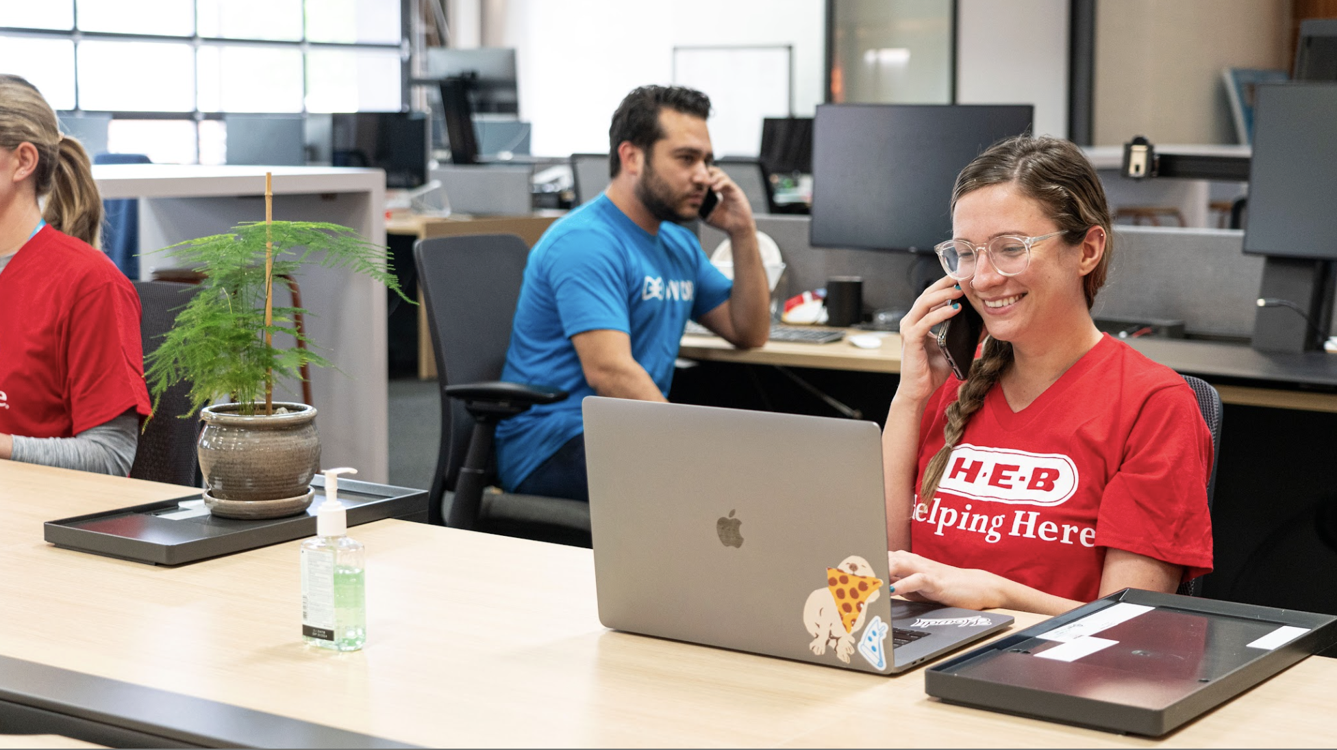 Office workers, a woman in a red shirt with H.E.B. logo smiling and talking on the phone, working on a laptop with dog and pizza stickers, in a modern office environment.
