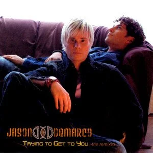 Two people sitting on a couch, one with short platinum blonde hair and the other with curly dark hair, in a relaxed setting.