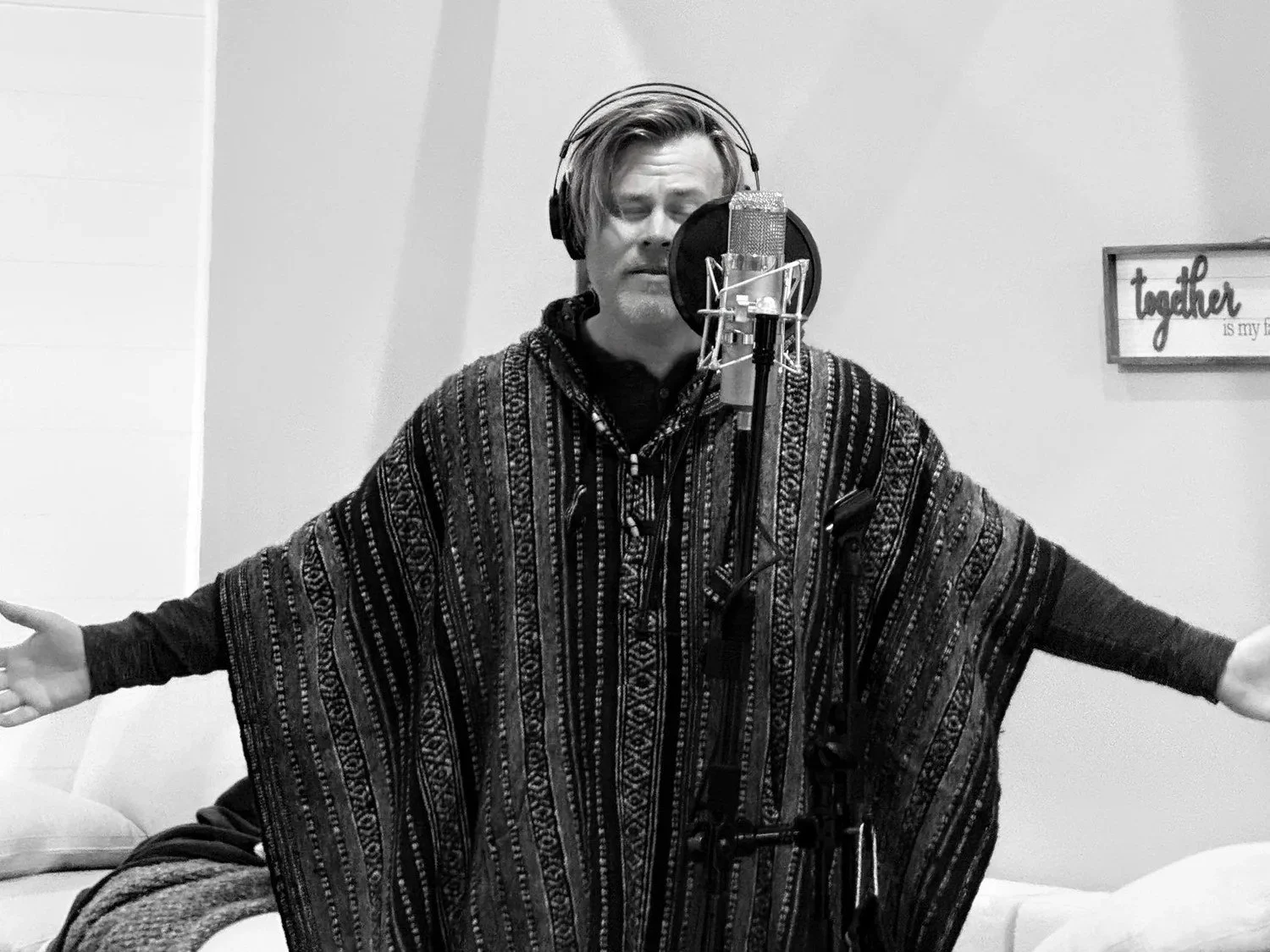 Man singing into a studio microphone with eyes closed, arms outstretched, wearing a patterned poncho and headphones, in a recording studio with a framed sign that reads 'together' on the wall.